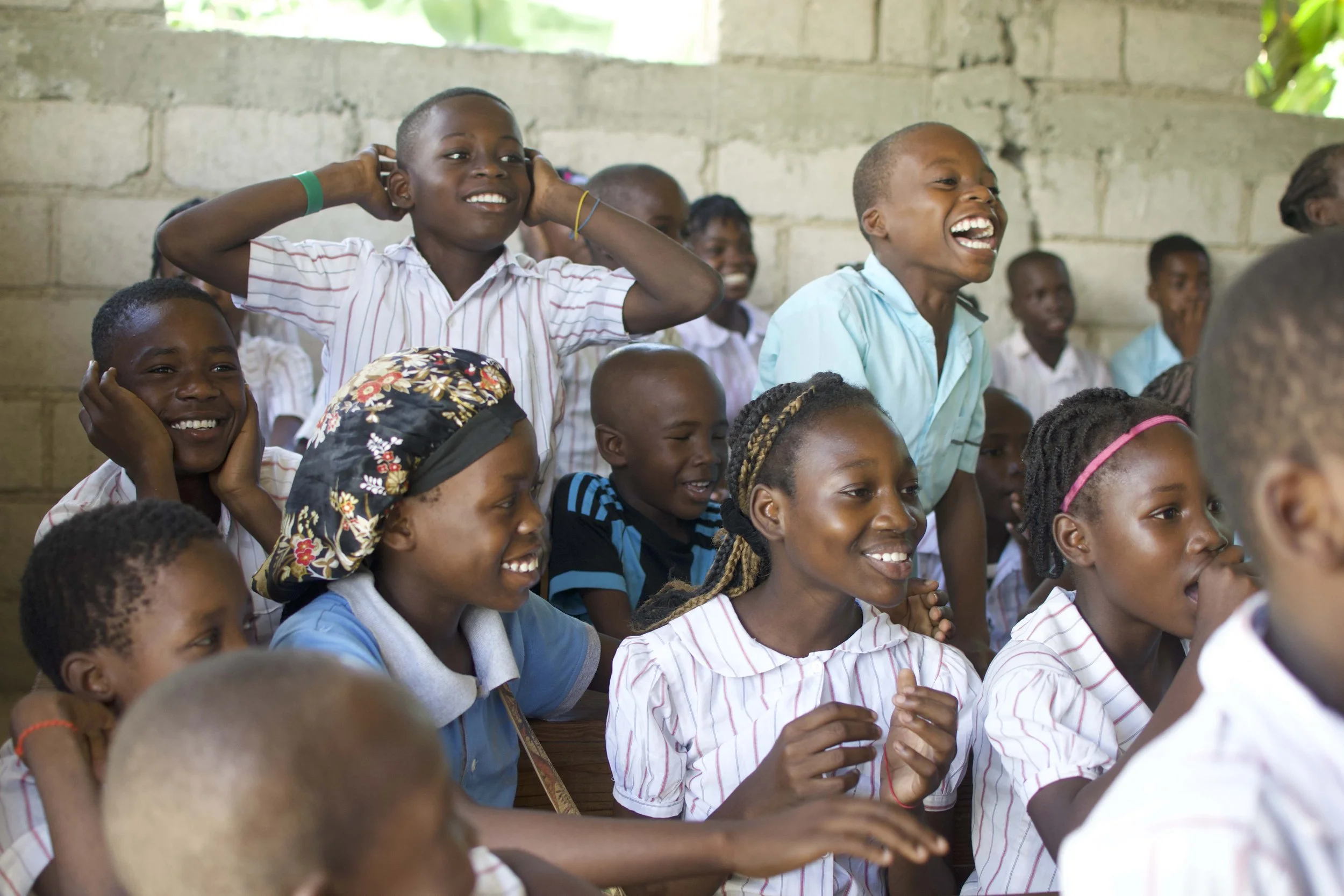 Group of smiling children in a classroom, some sitting and some standing, wearing school uniforms, with a concrete wall in the background.