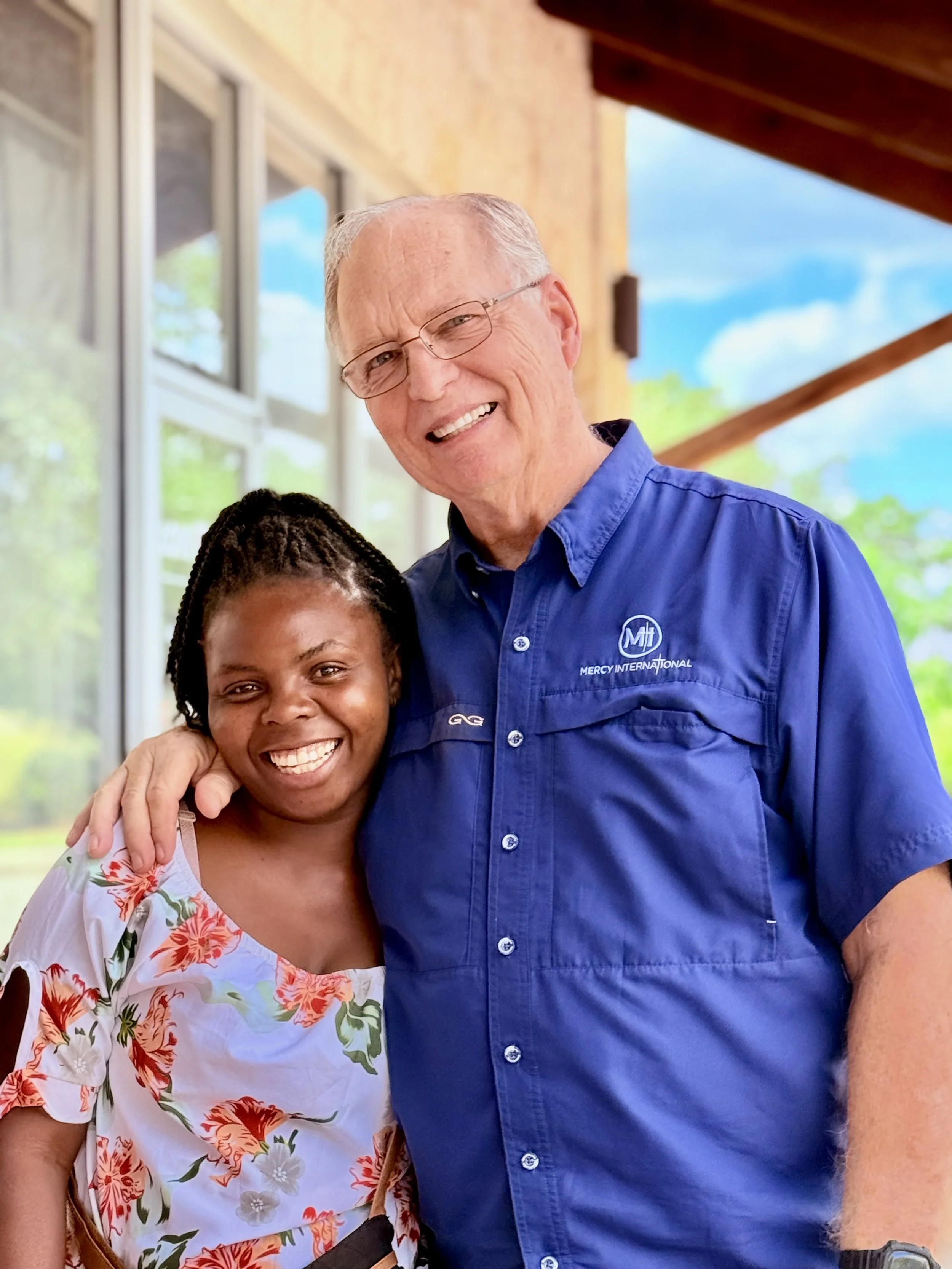 Two people smiling, one man and one woman, standing outside near a building with large windows and green trees in the background. The man has white hair, glasses, and is wearing a blue shirt with a logo that says 'Mercy International'. The woman has dark hair styled in braids and is wearing a floral dress.