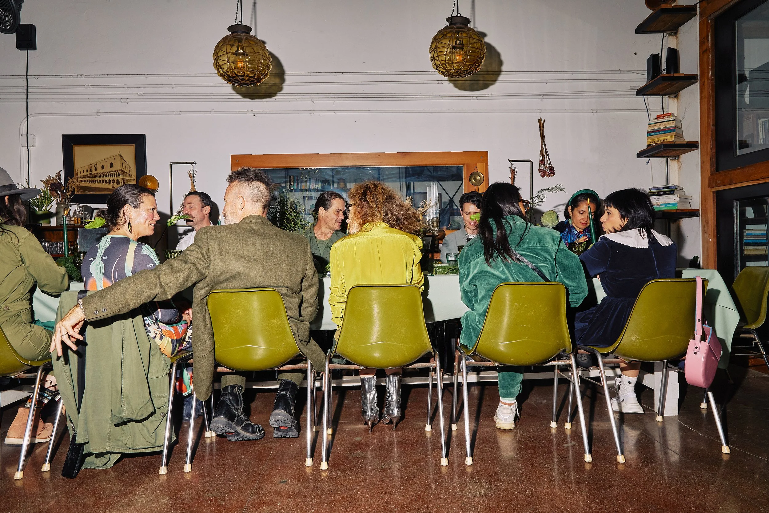 People sitting at a long table having a conversation in a cozy restaurant with white walls, warm lighting, and bookshelves.