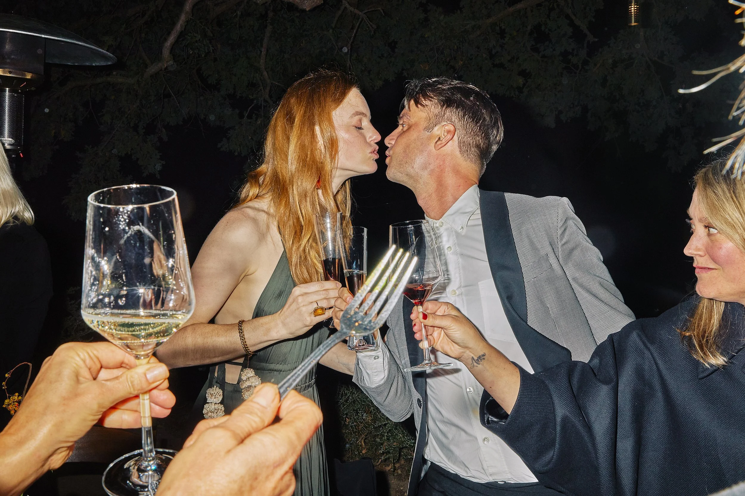 Editorial Wedding photography capturing a the bride and the groom sharing a kiss, holding glasses of wine at their wedding in Los Angeles, with other guests holding glasses in the foreground.