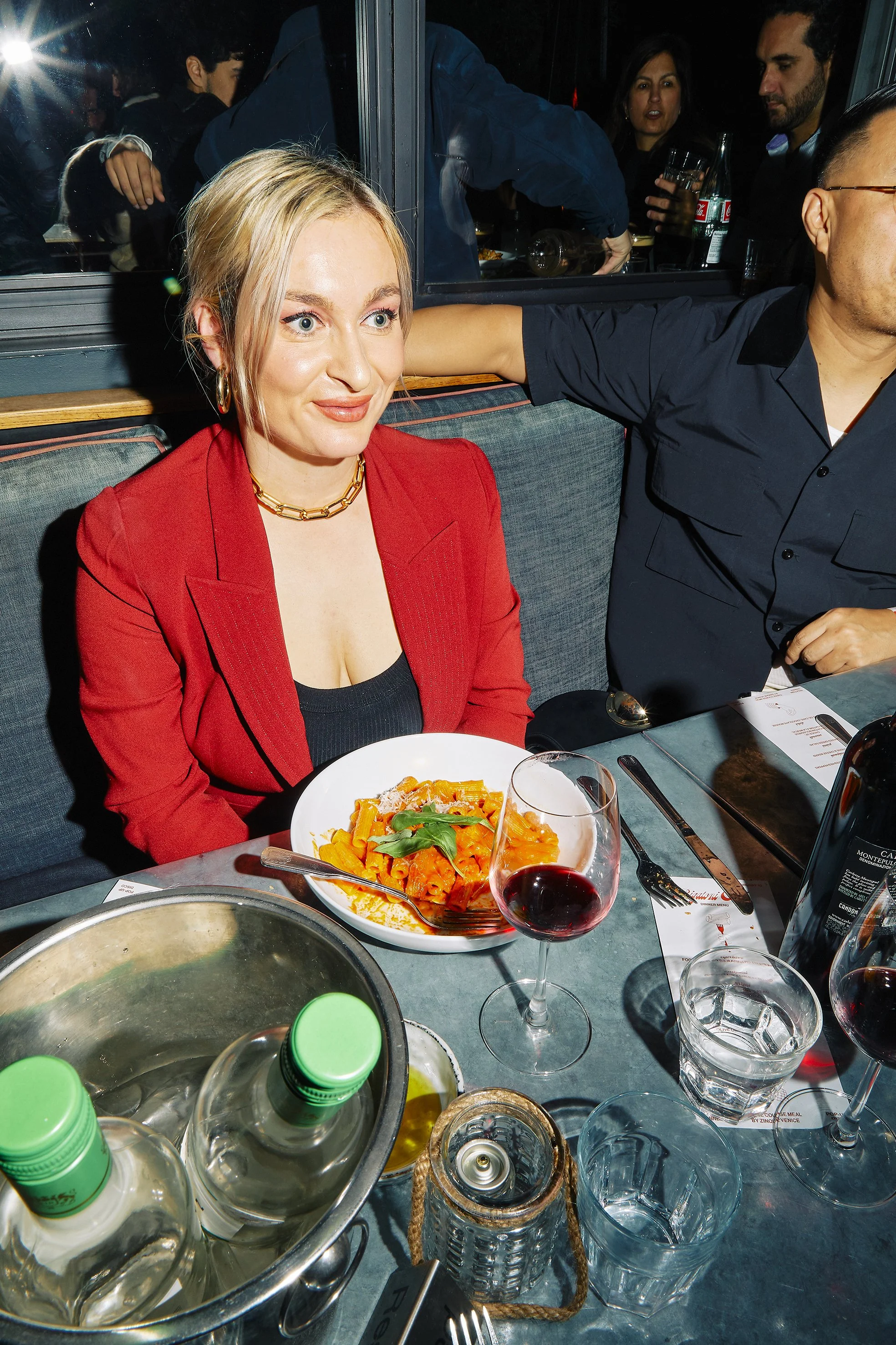 A woman with blonde hair wearing a red blazer and gold jewelry is sitting at a table with a plate of pasta, a glass of red wine, and water in front of her. Other people are visible in the background at a restaurant or bar.
