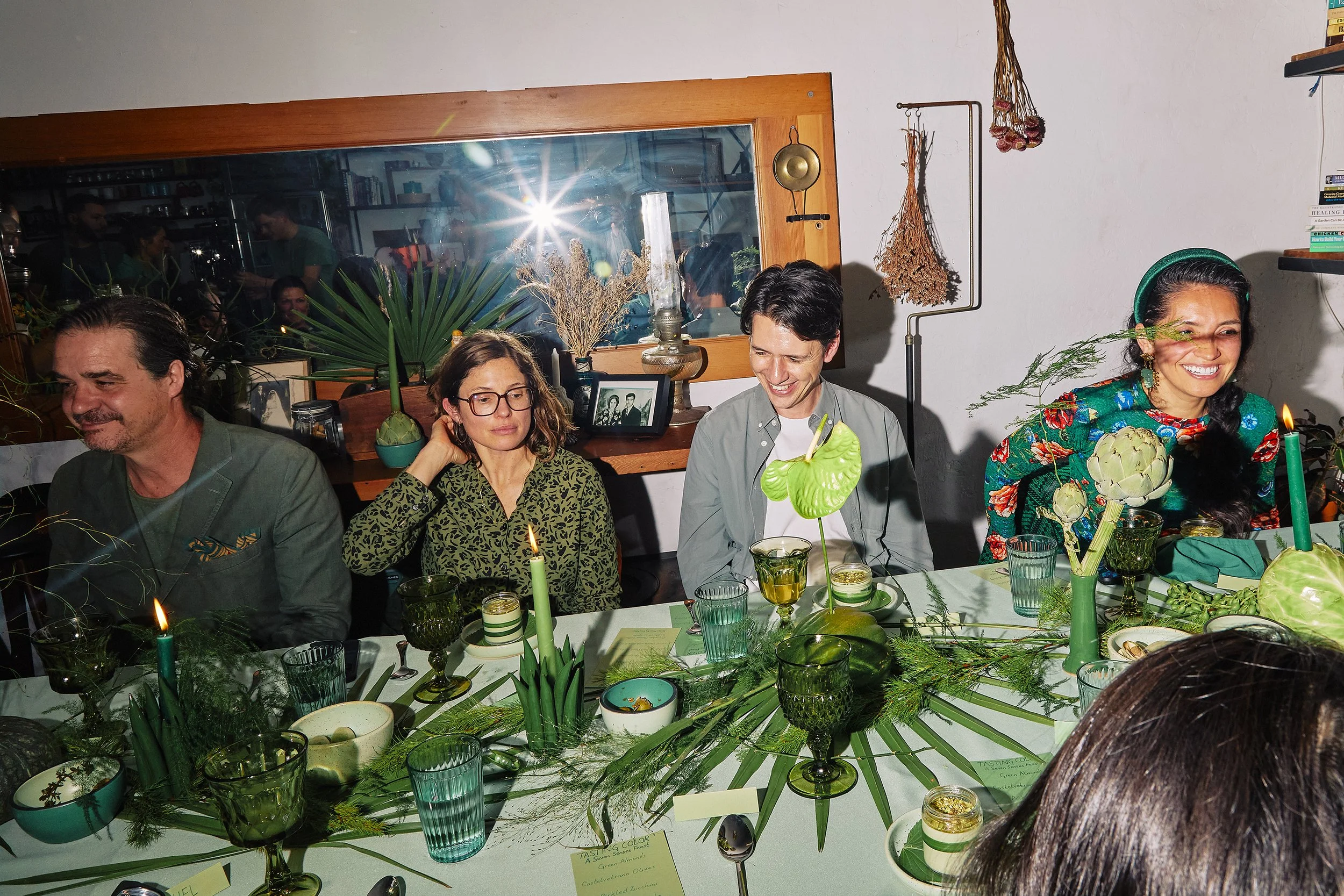 People sitting at a decorated dinner table with green and natural-themed decor, including candles, bowls, and leafy elements.