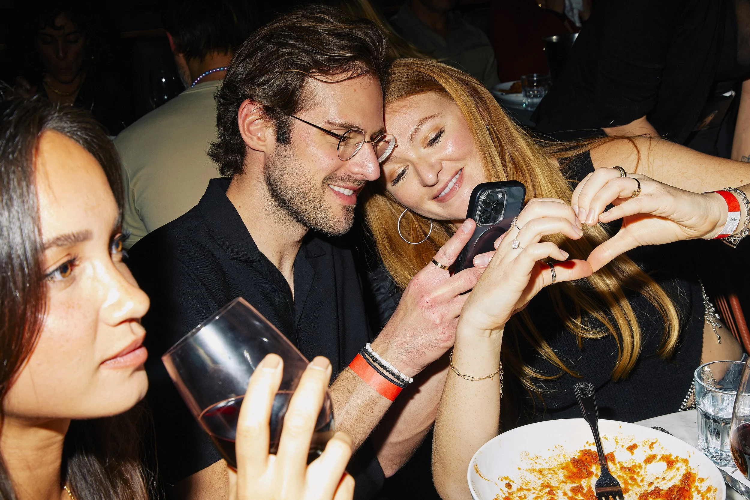 Three people sitting at a table, smiling and looking at a smartphone, with a woman showing heart shape with fingers. There are glasses of red wine and a plate with food in front of them.
