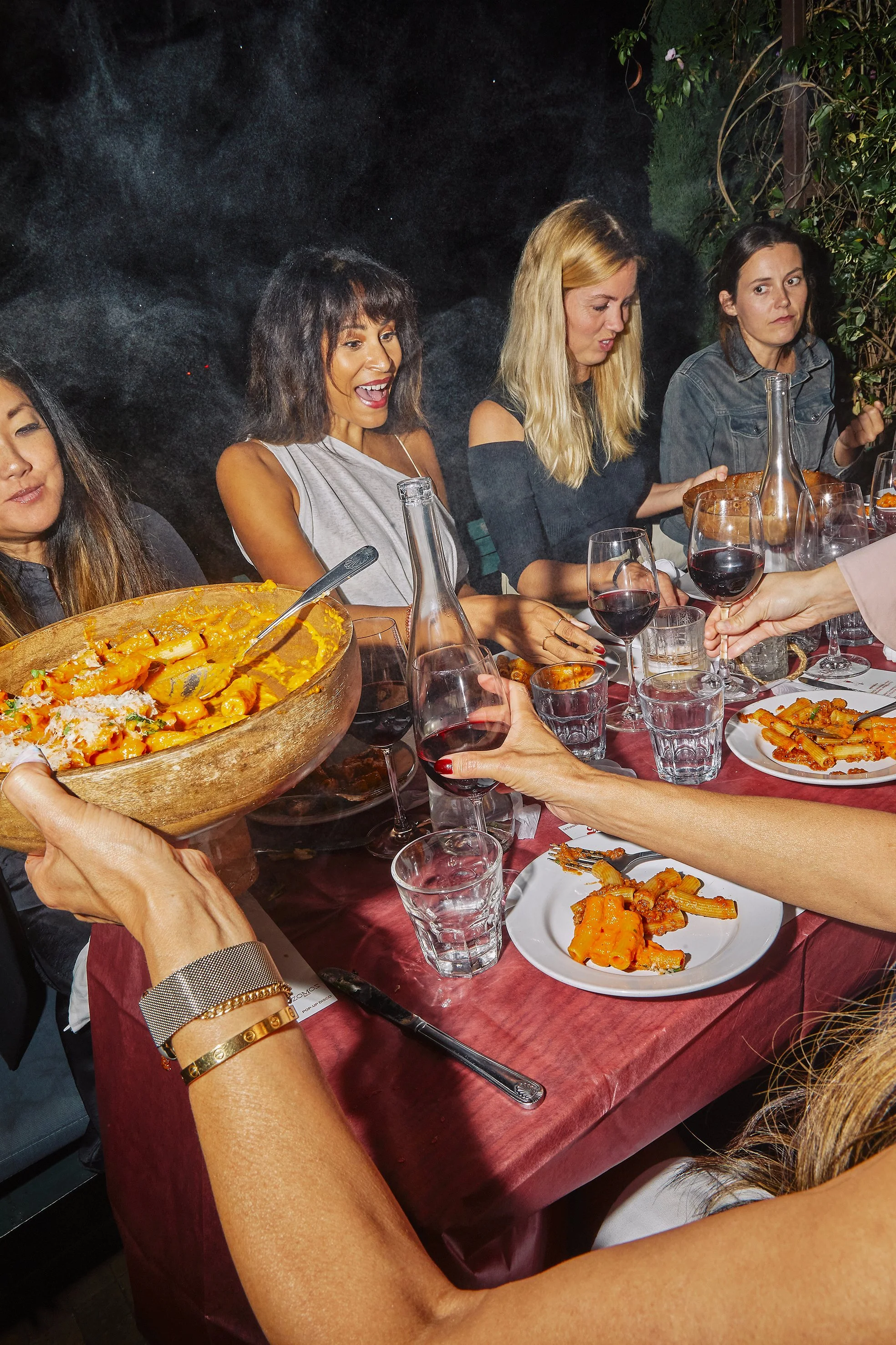 Editorial Event photography capturing a group of women seated at a table enjoying a meal and pouring red wine, with a large serving of pasta being passed around at Zinqué in Venice Los Angeles for Rigatoni Club.