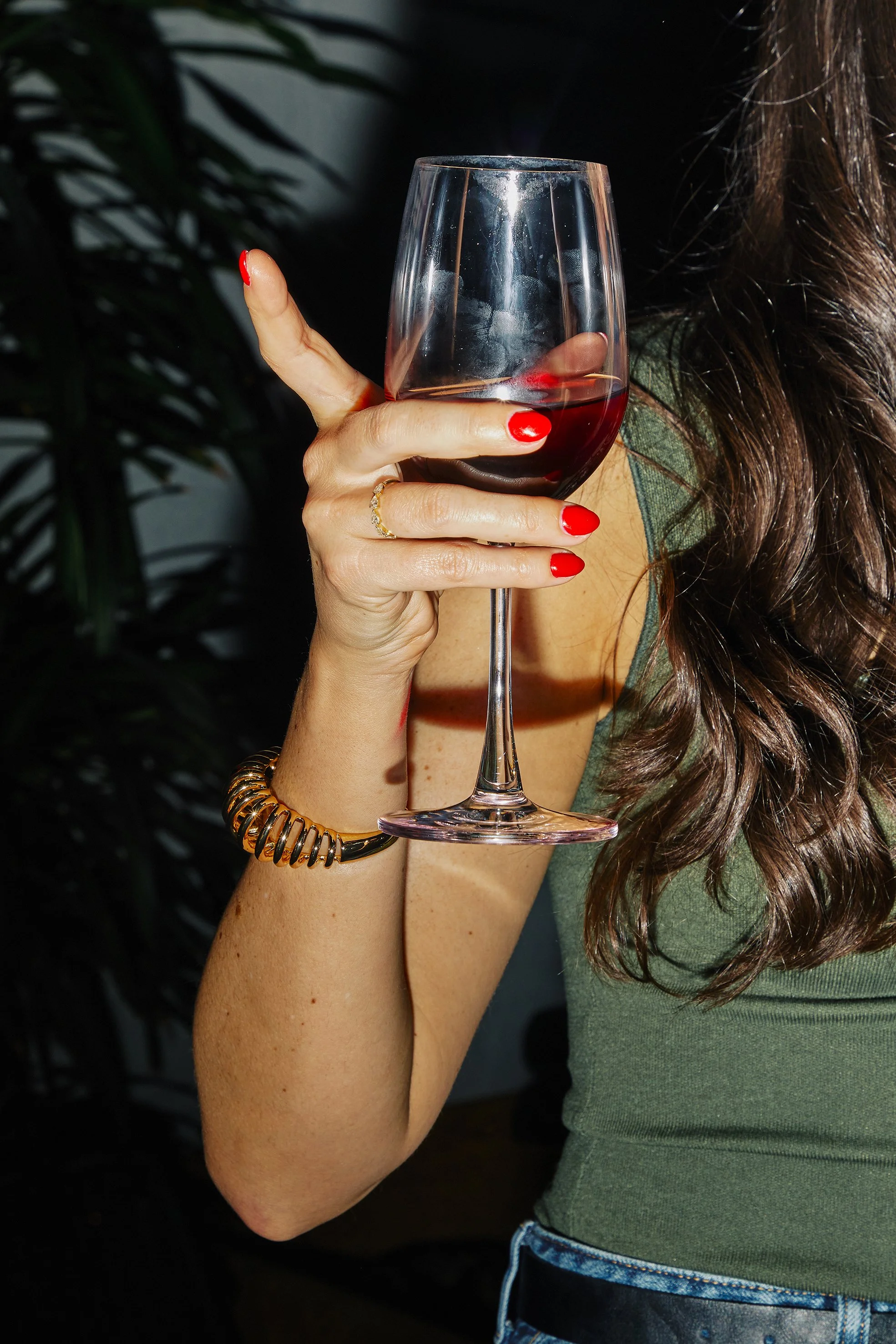 A woman holding a glass of red wine, with red painted nails, wearing a green top, a gold bracelet, and a gold ring.