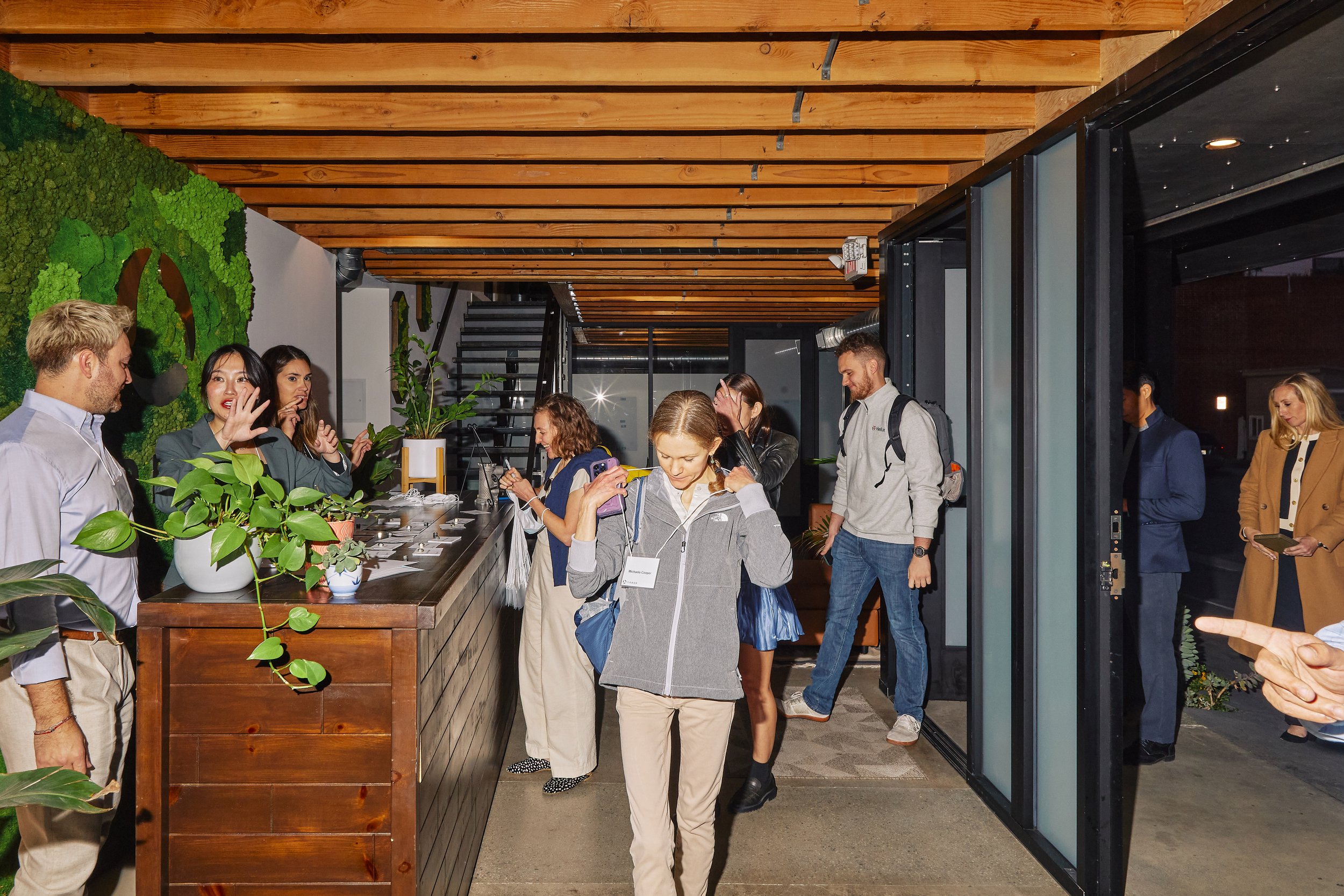 People checking in or registering at a reception desk inside a modern, warmly-lit office or hotel lobby with wooden ceiling and green plant decor.