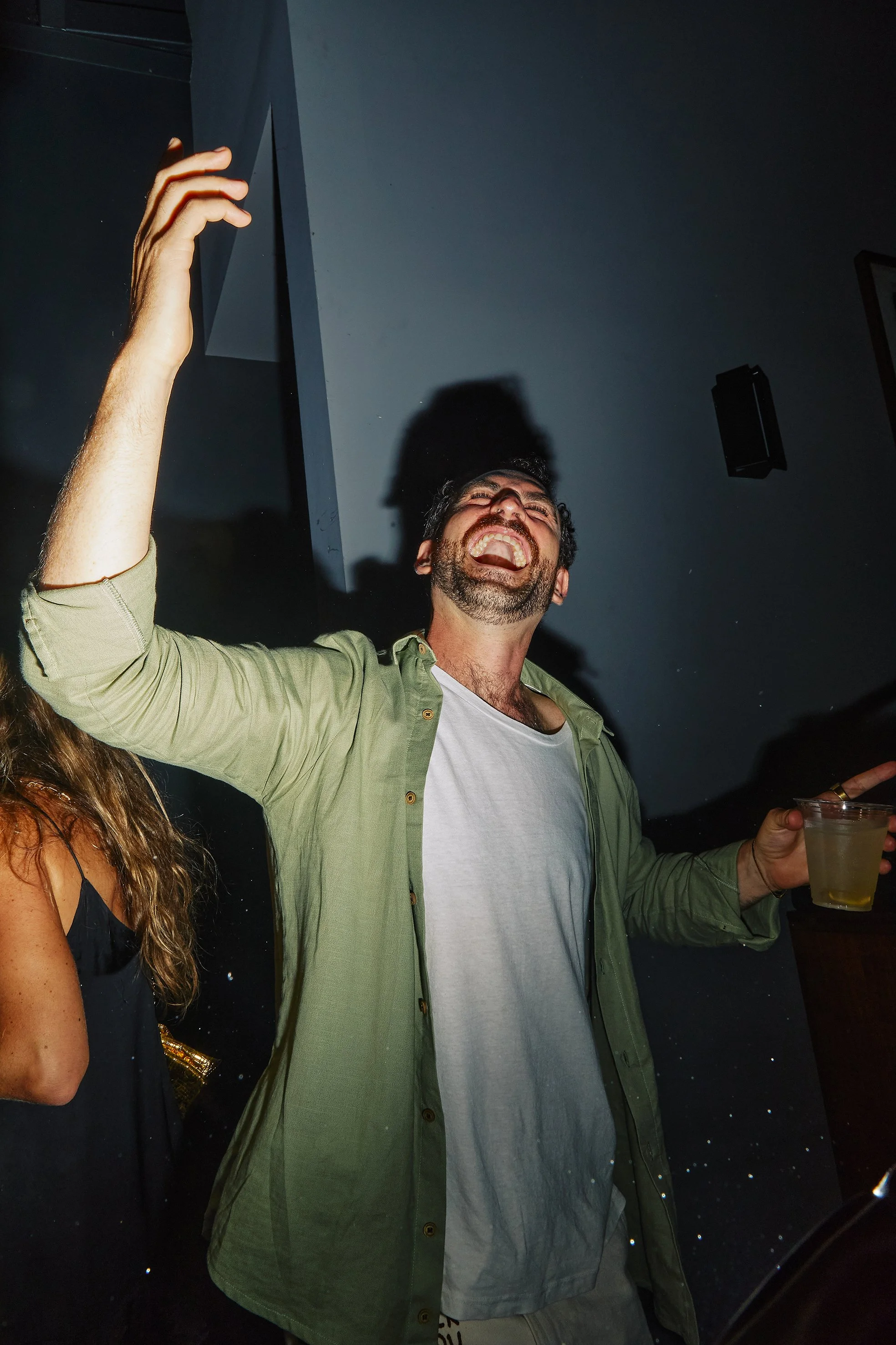 Man smiling and laughing, holding a drink in his right hand, at a party or social gathering.