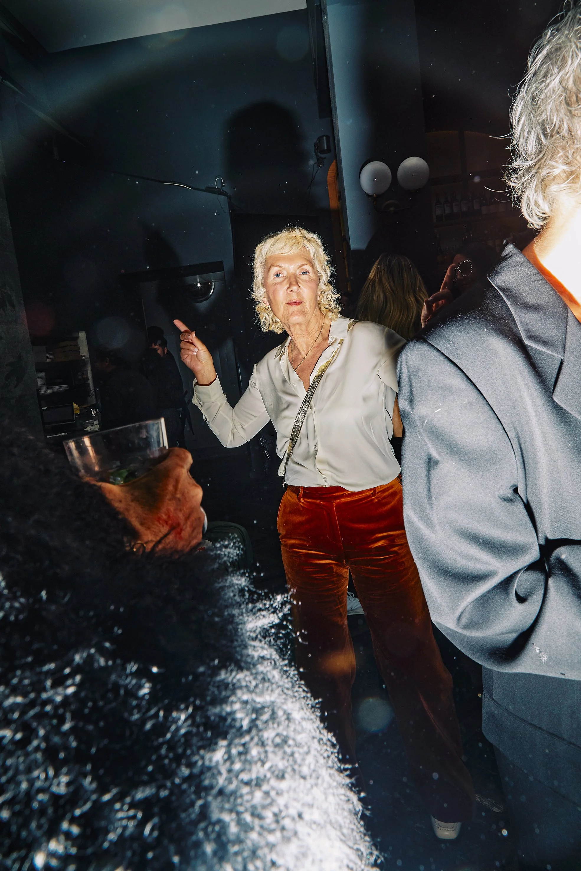 An elderly woman with curly gray hair gesturing with her index finger in a lively indoor setting among people, some of whom are partially visible. The scene appears to be at a social gathering or party.