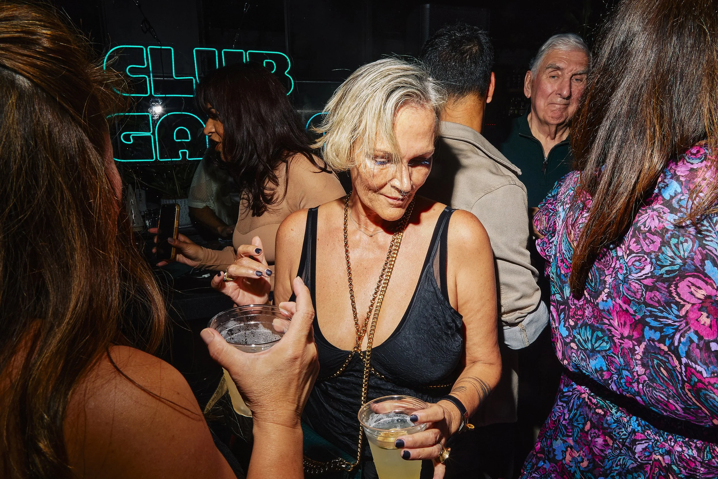 Group of people dancing and socializing in a club with a neon sign in the background that reads 'Club'.