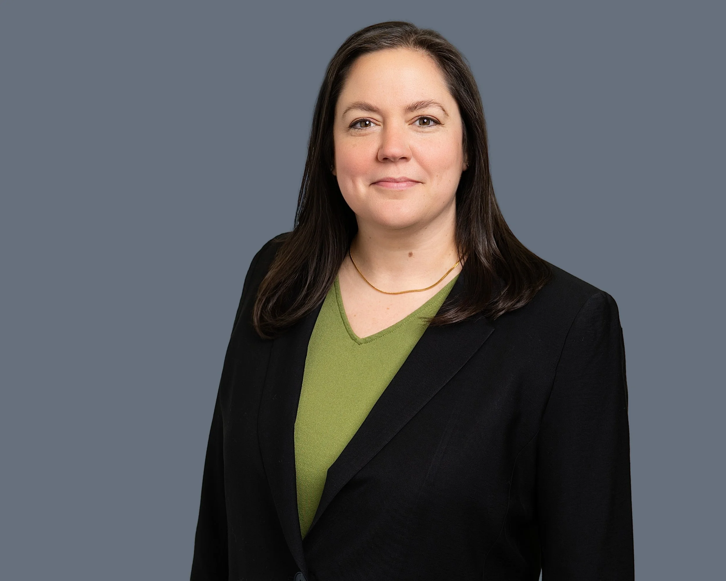 A woman with shoulder-length dark brown hair, wearing a black blazer over a green top, standing against a gray background.