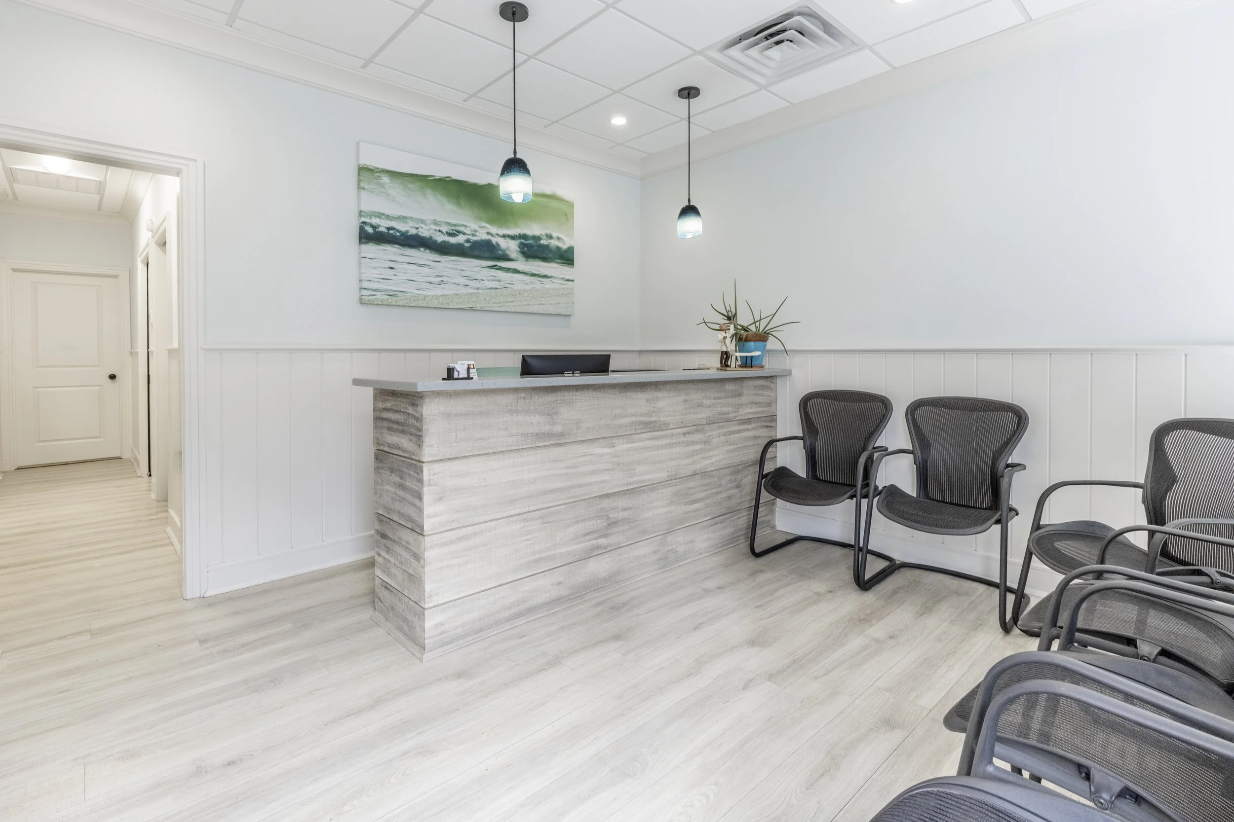 Empty reception area with a light-colored wooden desk, black mesh chairs, a plant, and a beach-themed painting of waves on the wall.