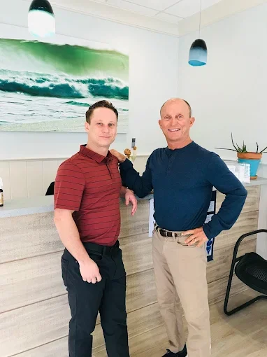 Two men standing inside a room with a surfboard wall art, one wearing a red striped polo shirt and the other in a navy blue sweater and khaki pants, smiling at the camera.