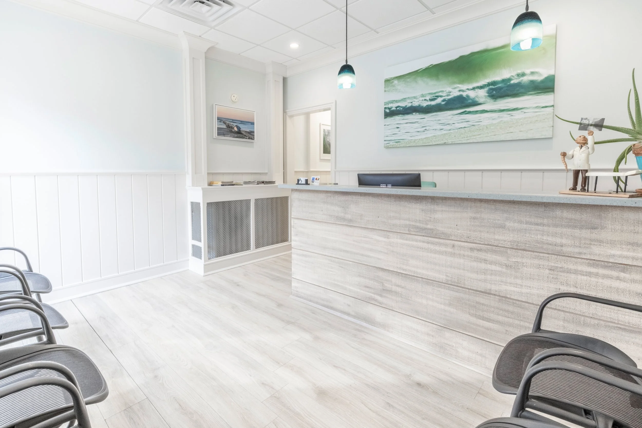 A clean, modern reception area with light-colored floors, a white desk, and beach-themed artwork on the walls. Several chairs are arranged along the walls for visitors.