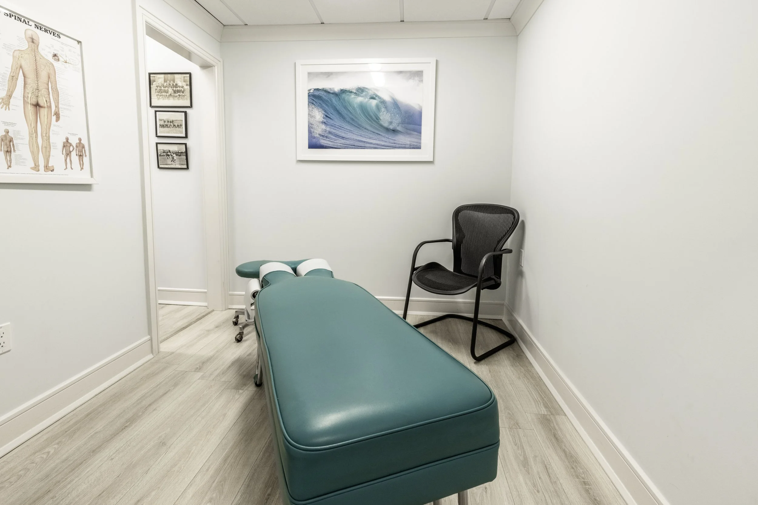 Medical examination room with a green examination table, a black chair, framed artwork of ocean waves and a world map, and a wall diagram of human nerves.