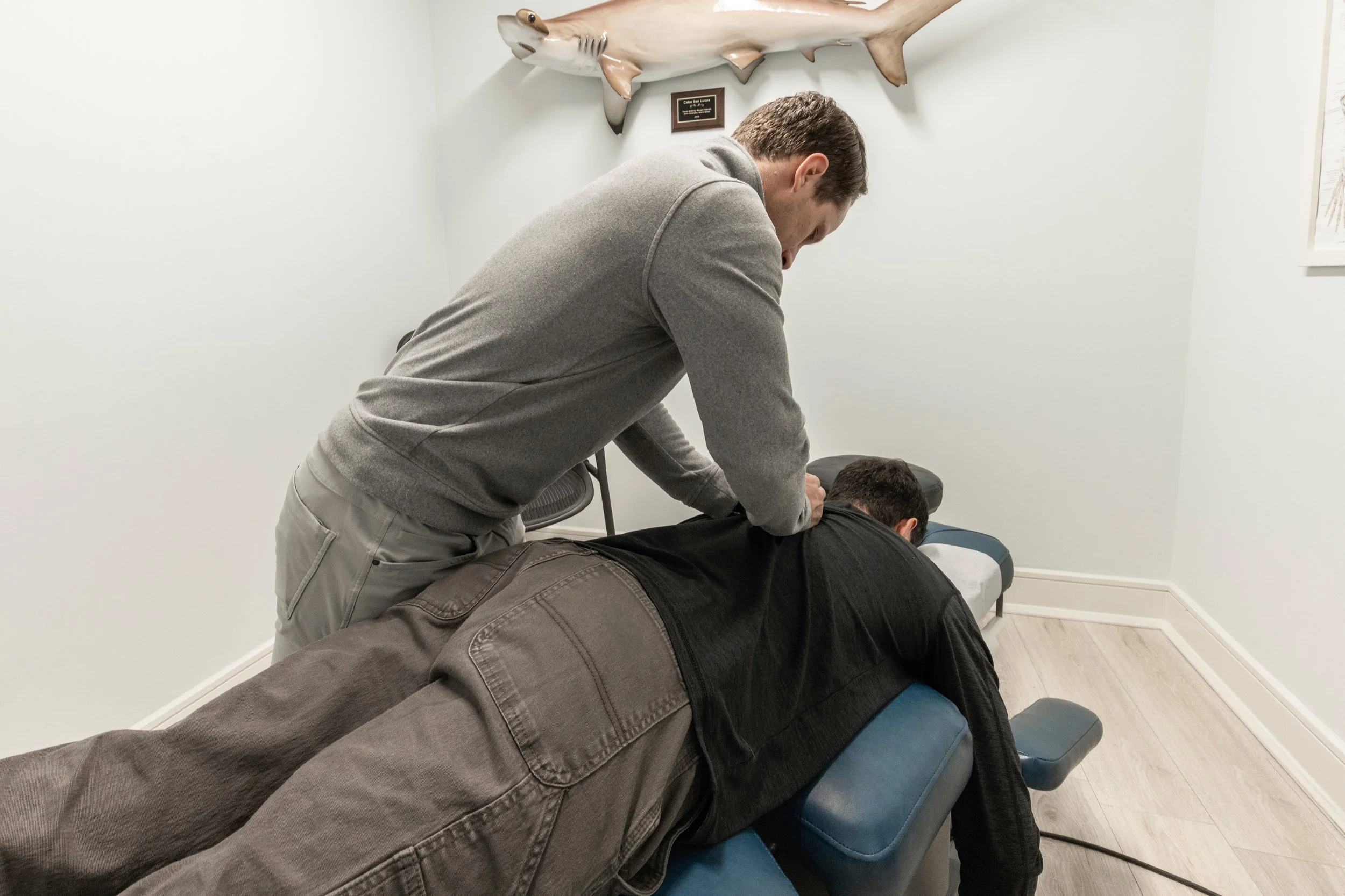 A chiropractor performs a neck adjustment on a patient lying face down on an adjustment table in a clinic room. A mounted fish sculpture is visible on the wall behind.