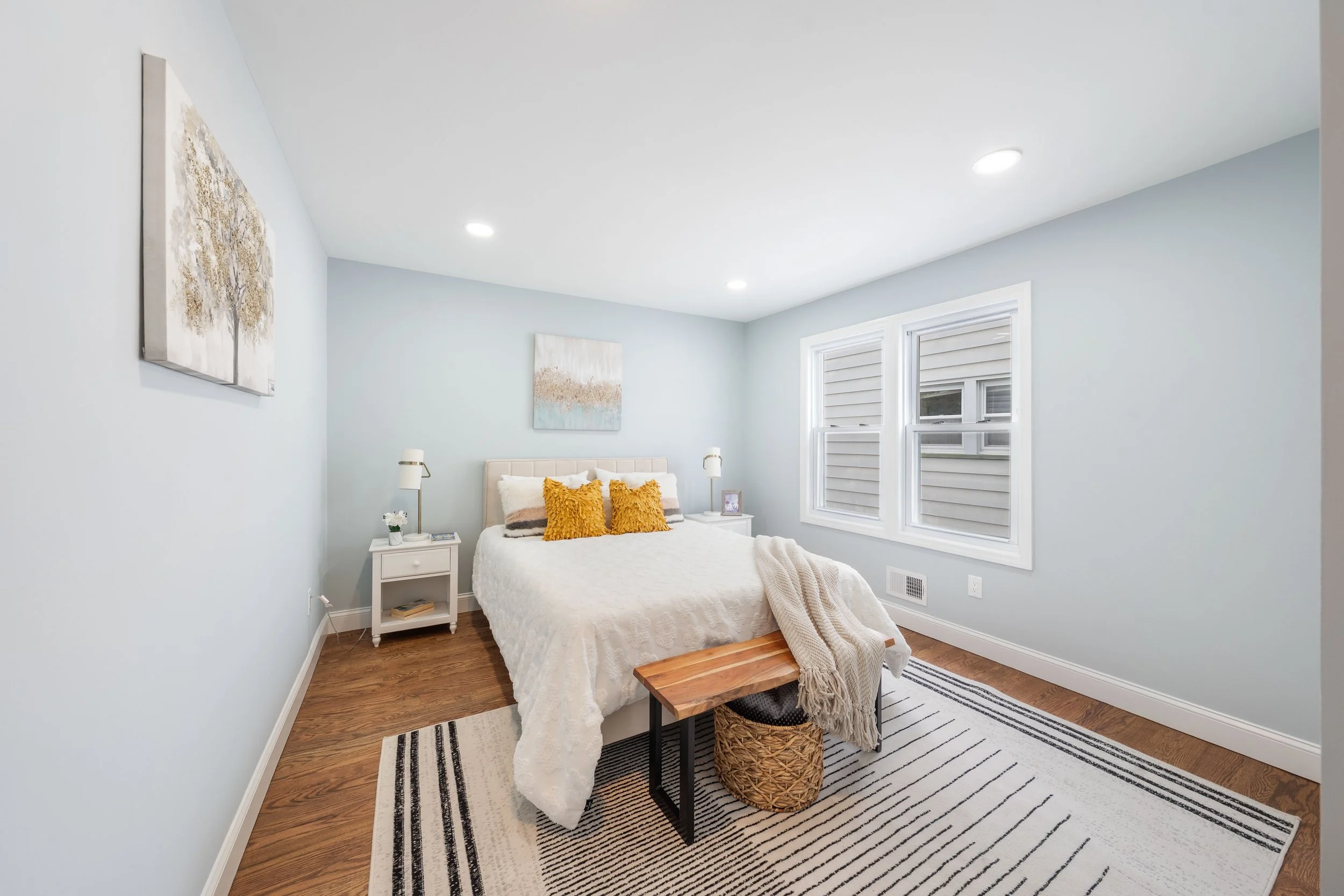 A bedroom with light blue walls, a white bed with orange throw pillows, two nightstands with lamps, and a striped rug on a wooden floor.
