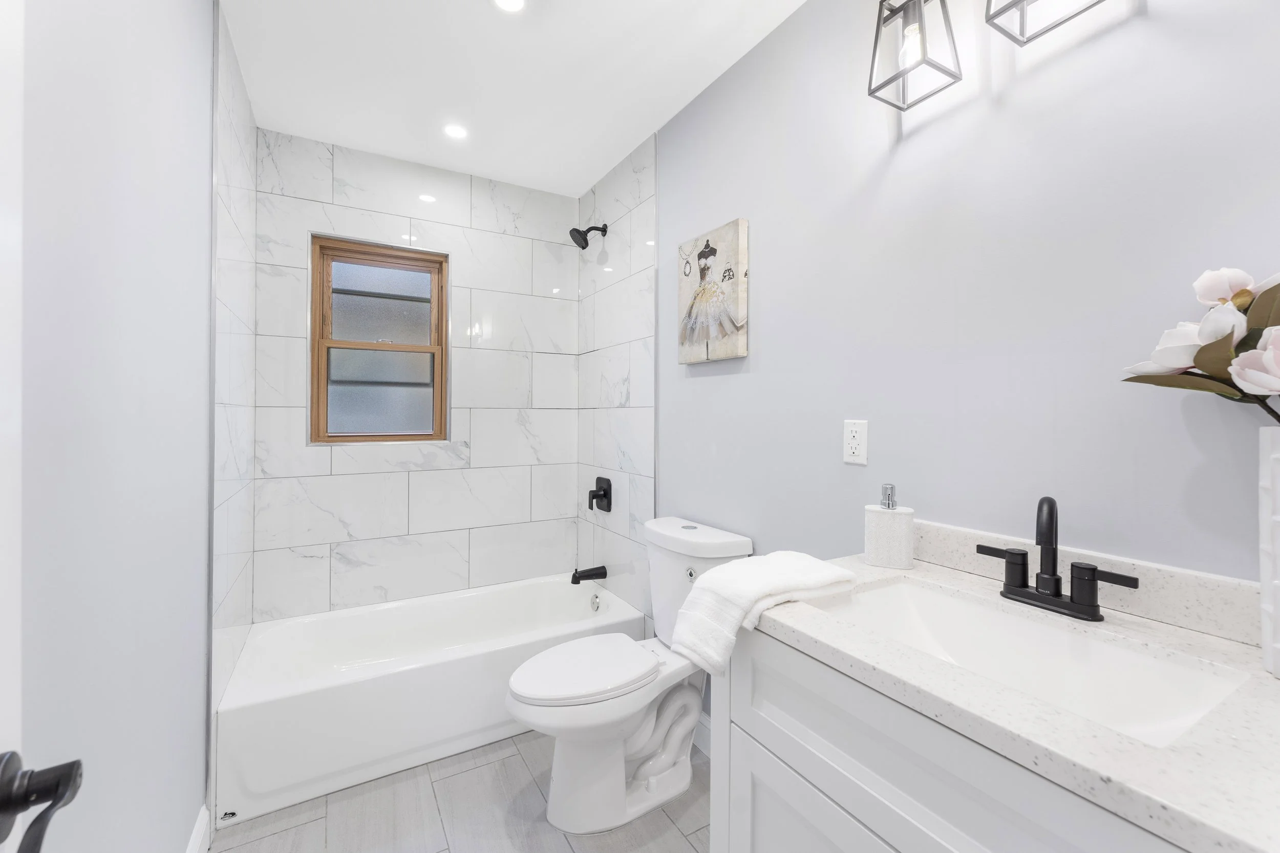 Modern bathroom with a white bathtub, toilet, and sink area featuring a black faucet. The wall behind the bathtub is tiled with white marble tiles. There is a small rectangular window with wood trim above the bathtub. Wall art depicting a ballerina i