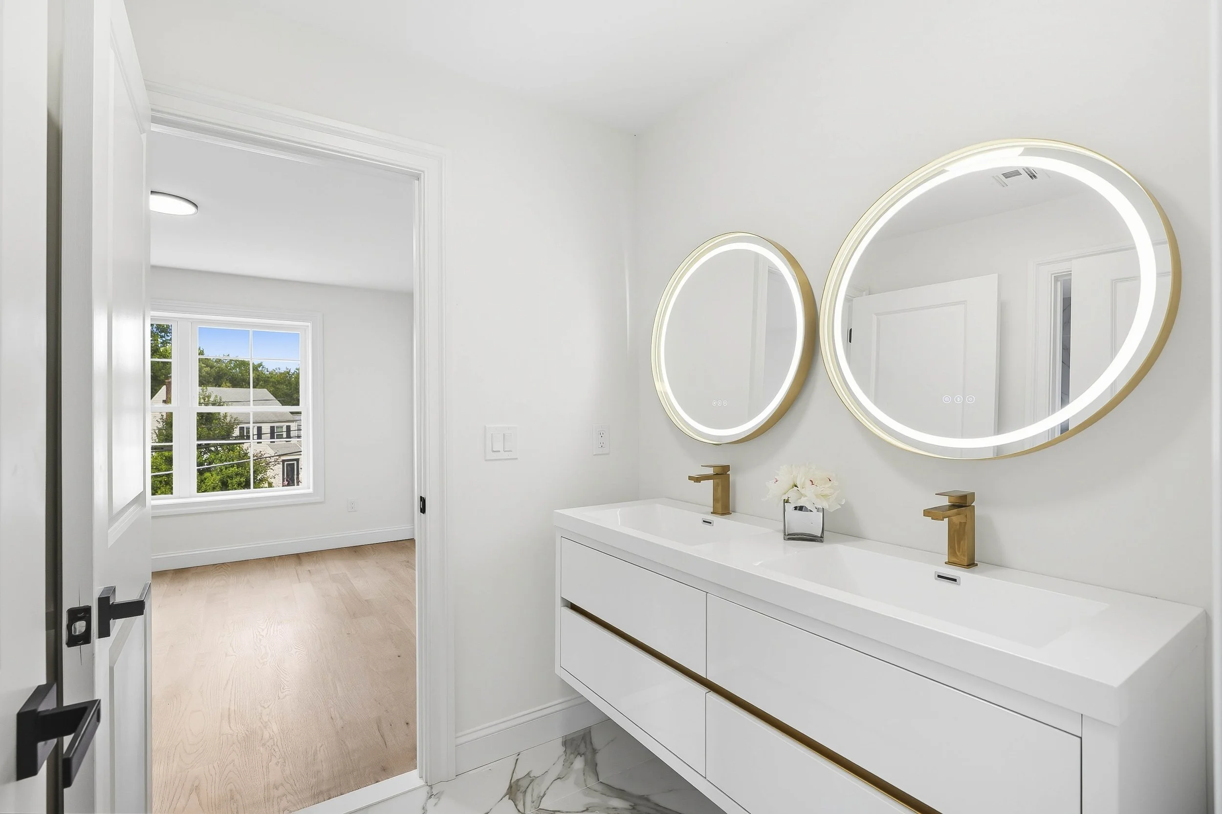 Modern bathroom with double vanity, oval illuminated mirrors, gold fixtures, white cabinetry, marble floor, and window showing outdoor greenery.