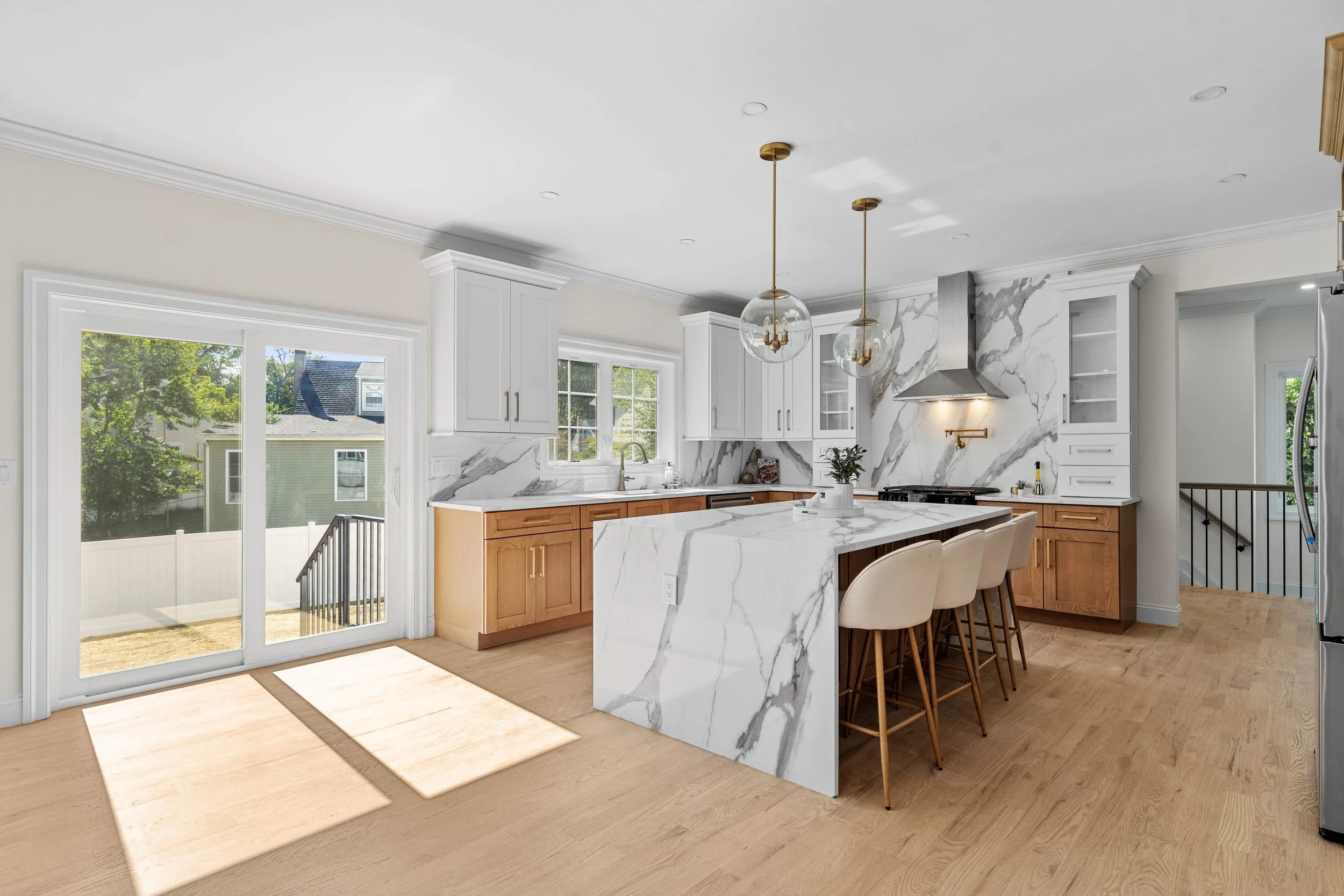 Bright, modern kitchen with white marble countertops and backsplash, natural wood cabinets, a large island with white chairs, stainless steel appliances, and a sliding glass door leading outside.