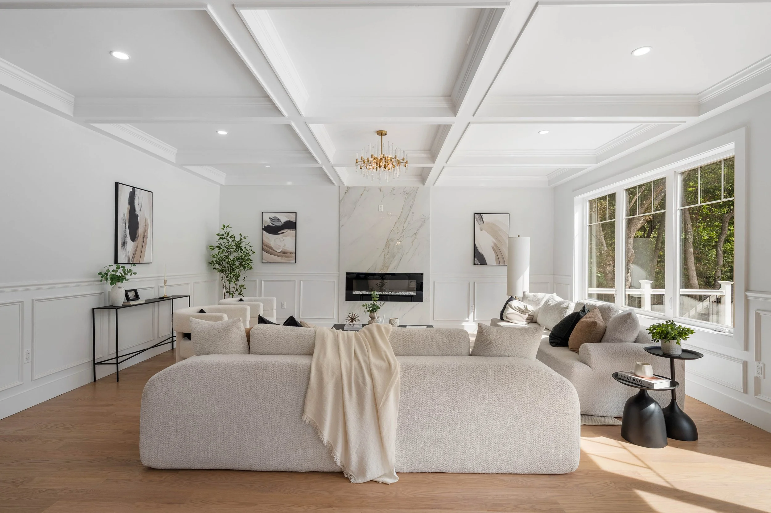 Bright, modern living room with white walls, large windows, and a beige sectional sofa. Artworks are hung on the walls, and a chandelier hangs from a coffered ceiling. Wooden flooring and a few plants add warmth and greenery.