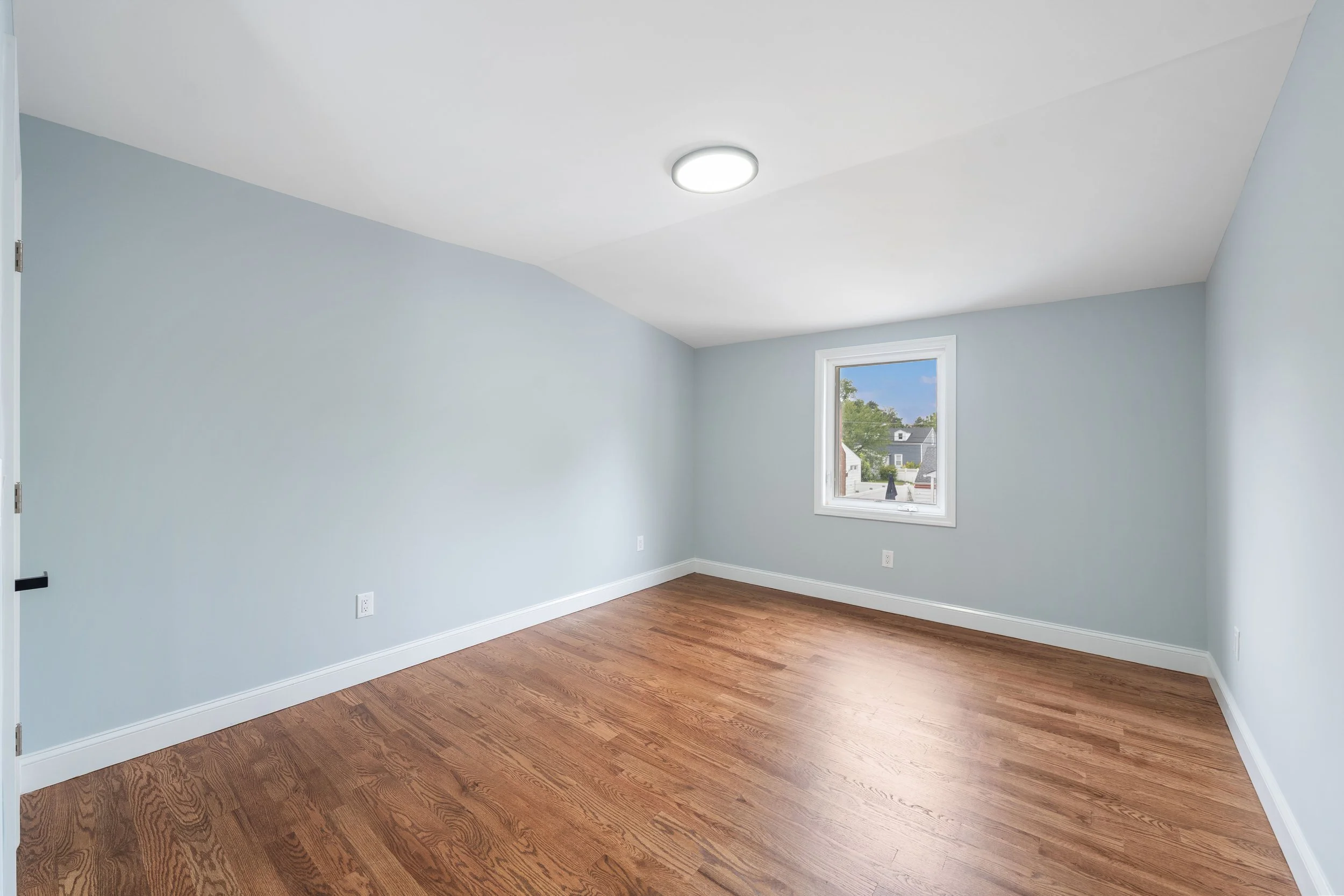 Empty bedroom with light blue walls, a window showing a neighborhood view, and hardwood floors.