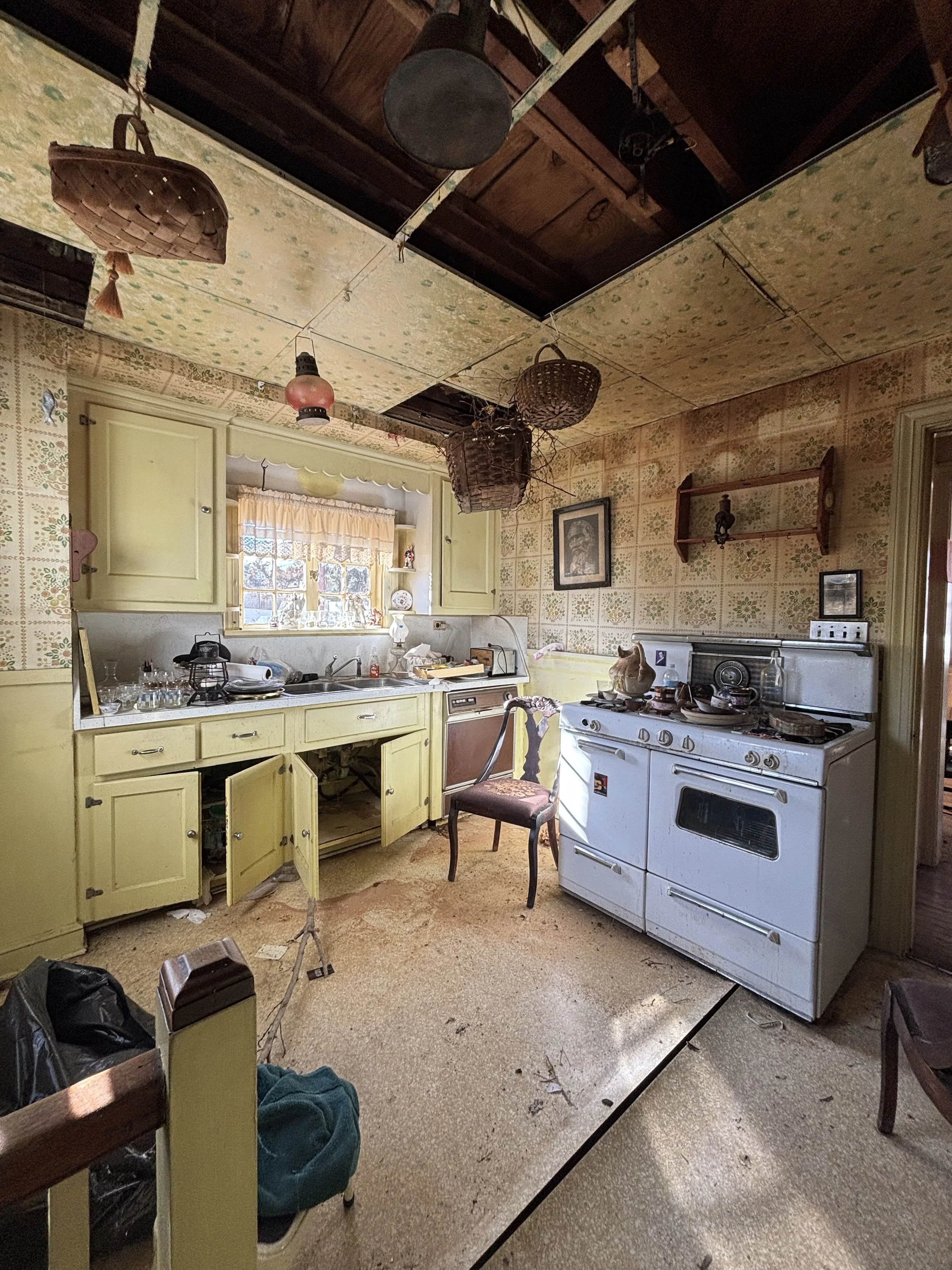 An old kitchen with yellow cabinets, a white stove, a chair, and a window with sunlight. The ceiling is deteriorated with hanging baskets and a lampshade, and the floor is dirty and stained.