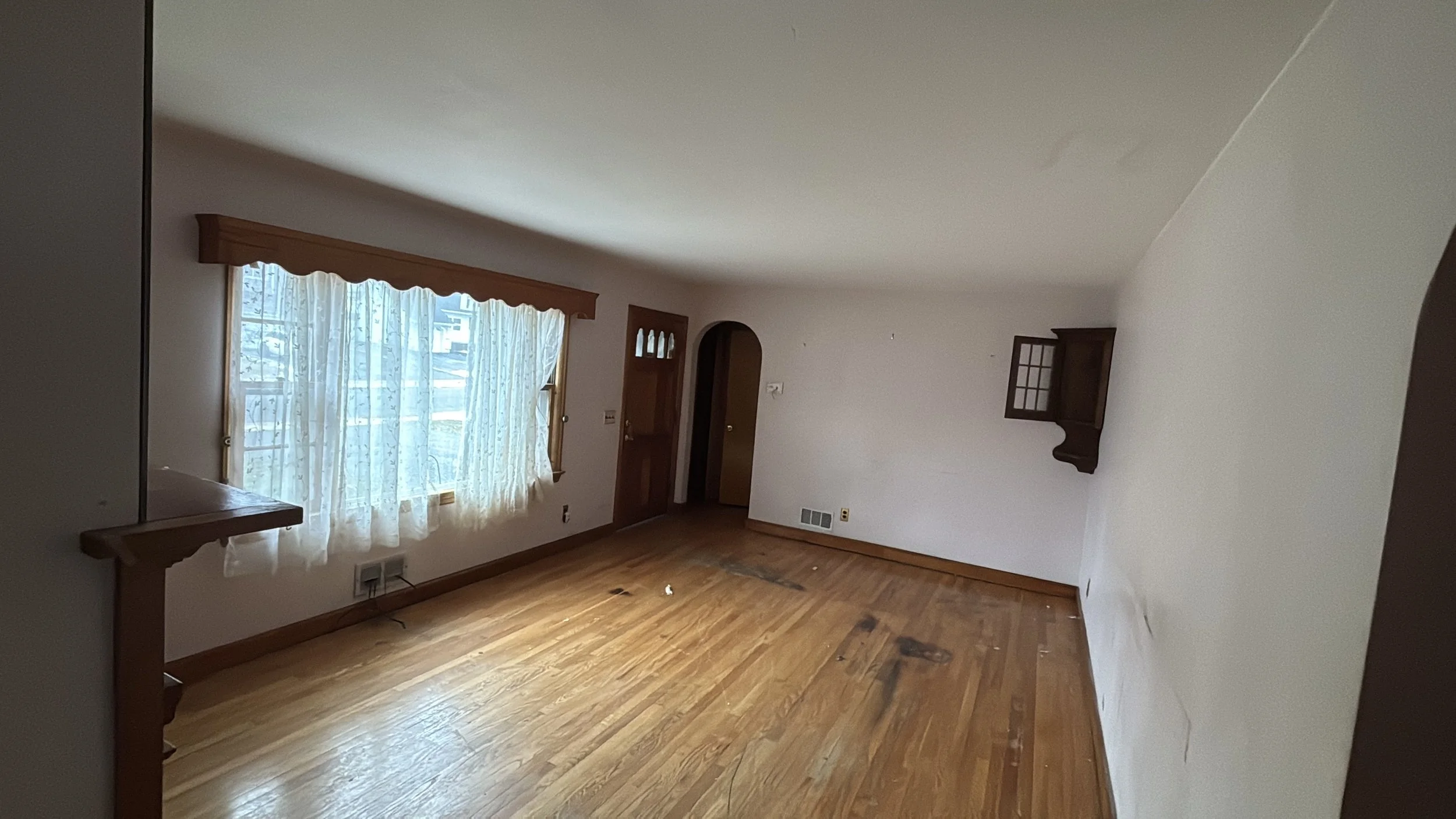 Empty living room with hardwood floors, a large window with curtains, and small wall-mounted cabinets.