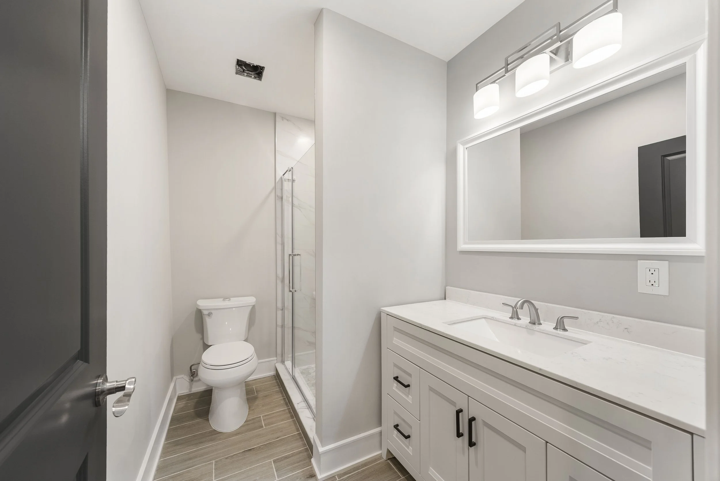 Modern bathroom with white vanity, marble countertop, large mirror with two lights, toilet, and glass shower enclosure, light-colored walls, wood-like floor tiles.