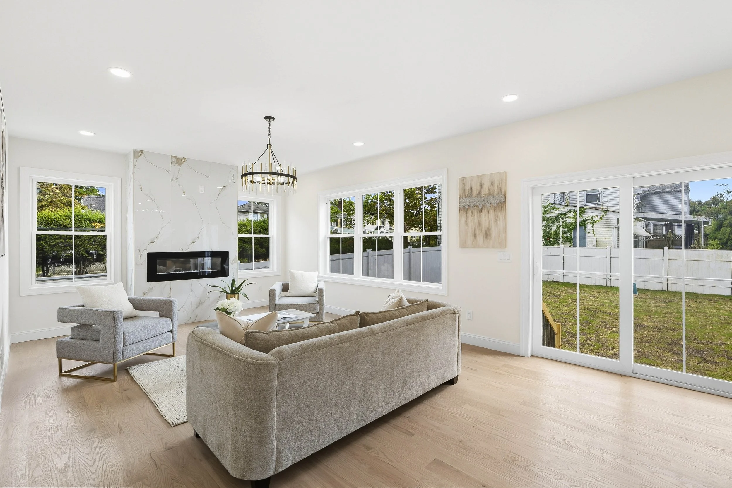 Bright living room with beige sofa, two grey armchairs, a white rug, a modern fireplace with marble surround, large windows, and a sliding glass door opening to a backyard.