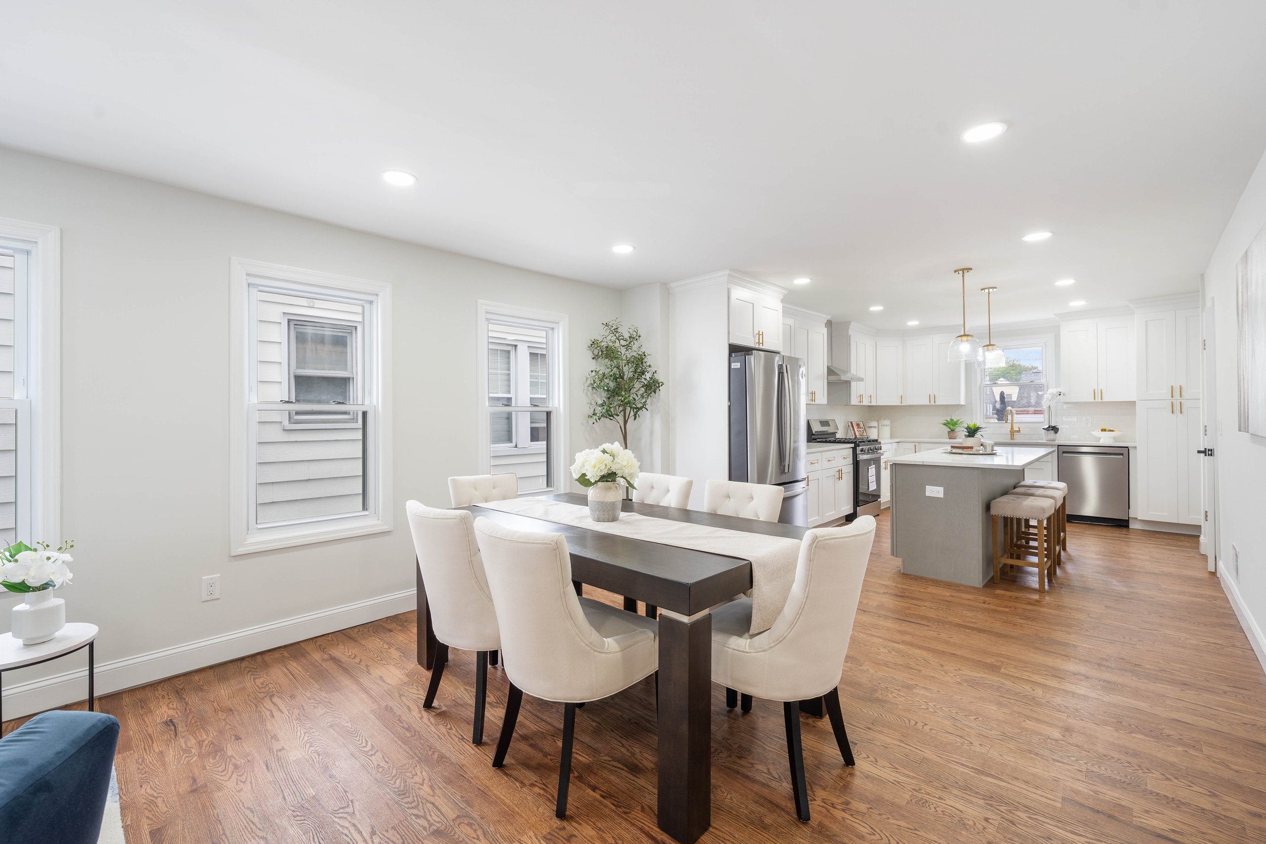 A spacious, modern kitchen and dining area with white walls, hardwood floors, a wooden table with six cream-colored chairs, and a kitchen with white cabinets, stainless steel appliances, a small island with barstools, and pendant lights.