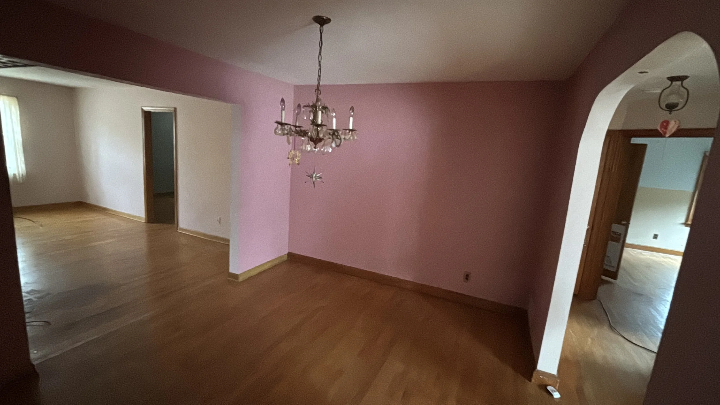 View of a living room with wooden floors, pink and white walls, a brass chandelier, and an archway leading to another room.