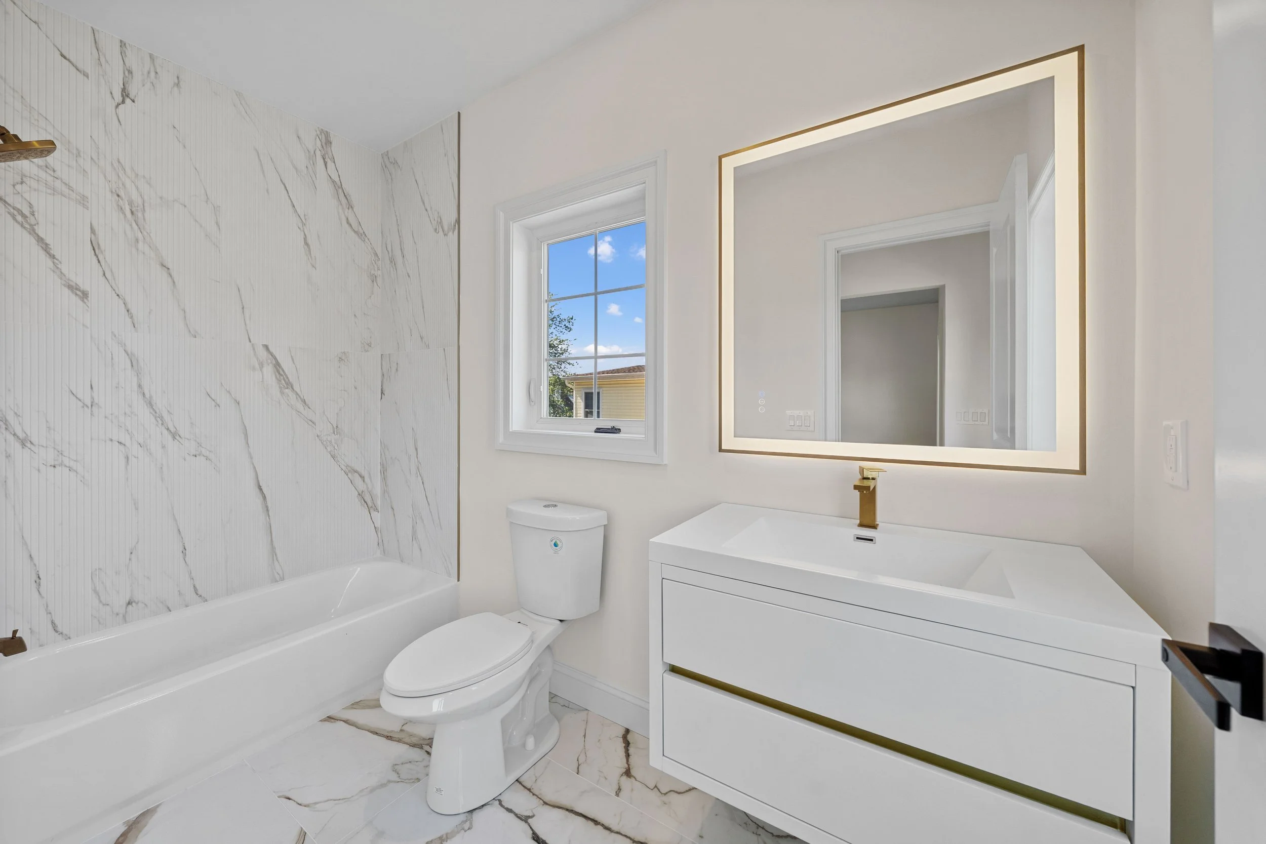 Modern bathroom with white marble floor and wall tiles, a white bathtub, a toilet, a white vanity with a large backlit mirror, and a window showing a blue sky and trees.