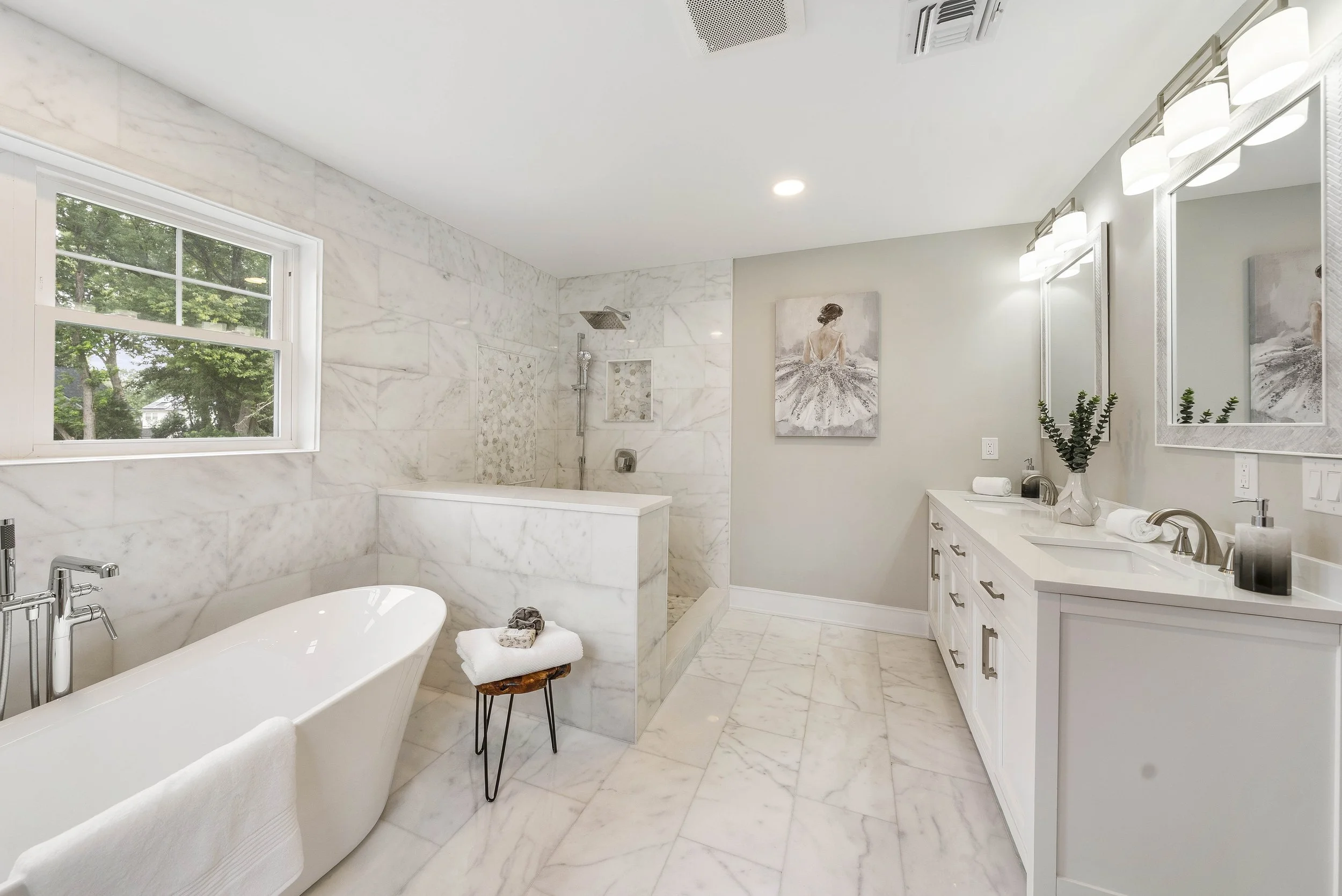 Modern white bathroom with a freestanding bathtub, a marble shower with a built-in niche, a white vanity with two mirrors, and artwork of a woman in a dress, with a window showing green trees outside.