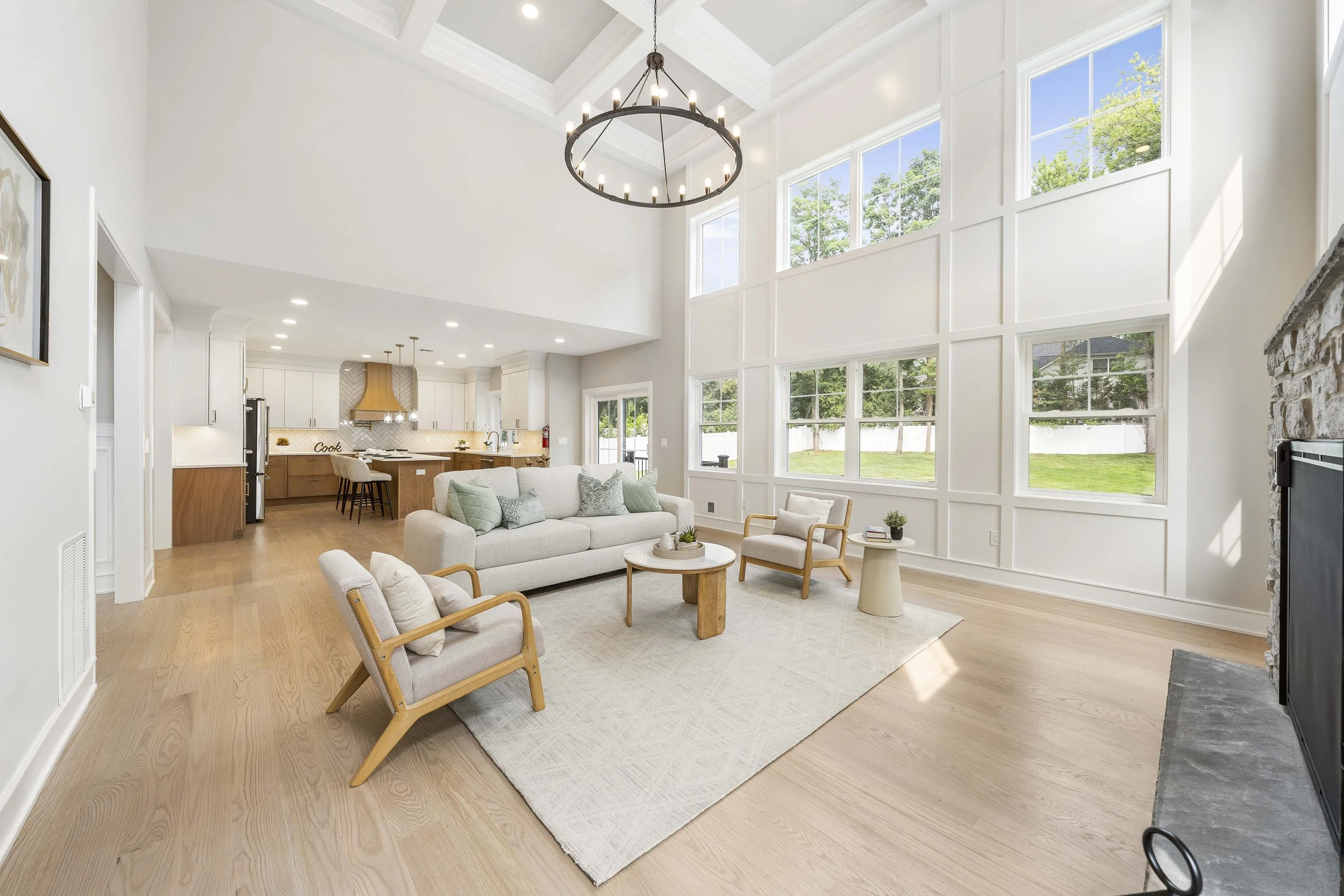 Spacious living room with large windows, white walls, and wood flooring. Contains a white sofa, two wooden armchairs, a small white side table, and a round coffee table, with a fireplace on the right. The open kitchen is visible in the background wit