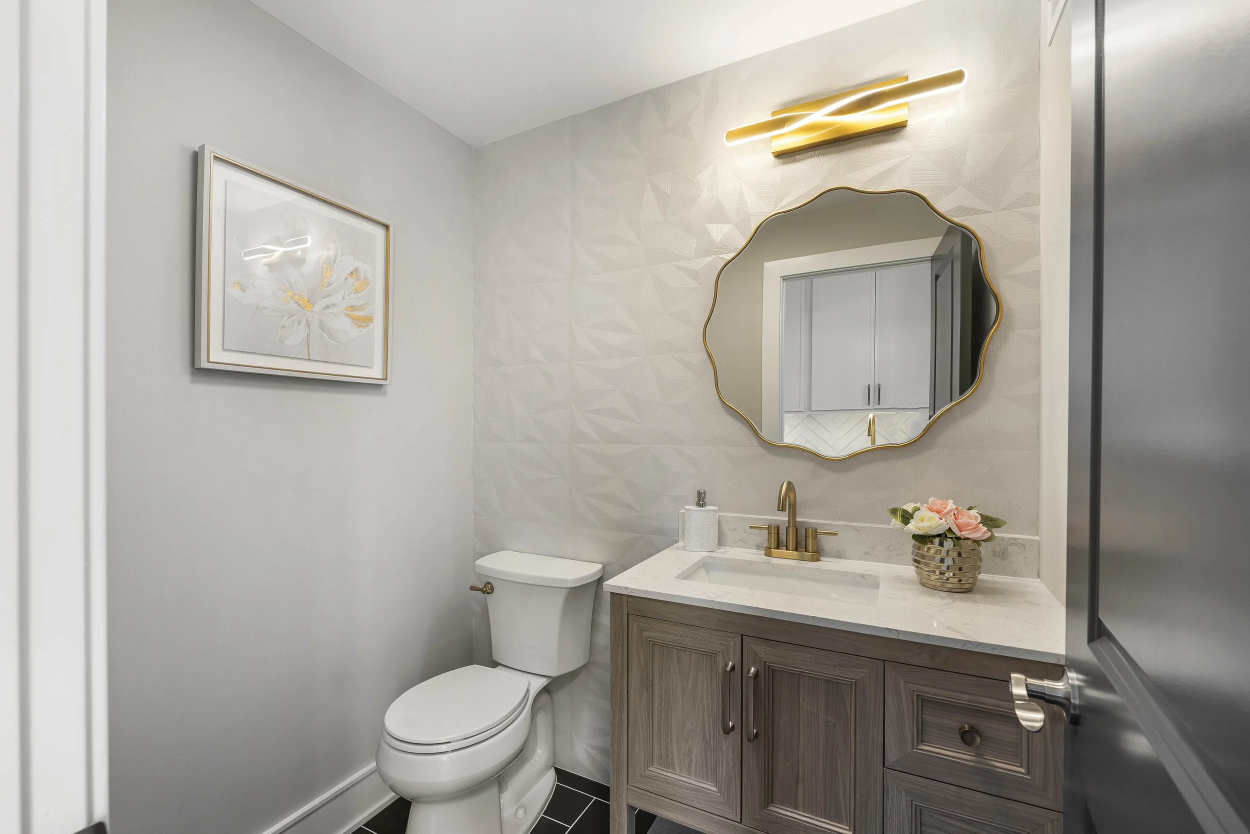Modern bathroom with a white toilet, wooden vanity with marble countertop, gold fixtures, decorative mirror, flower arrangement, wall art, and a light fixture