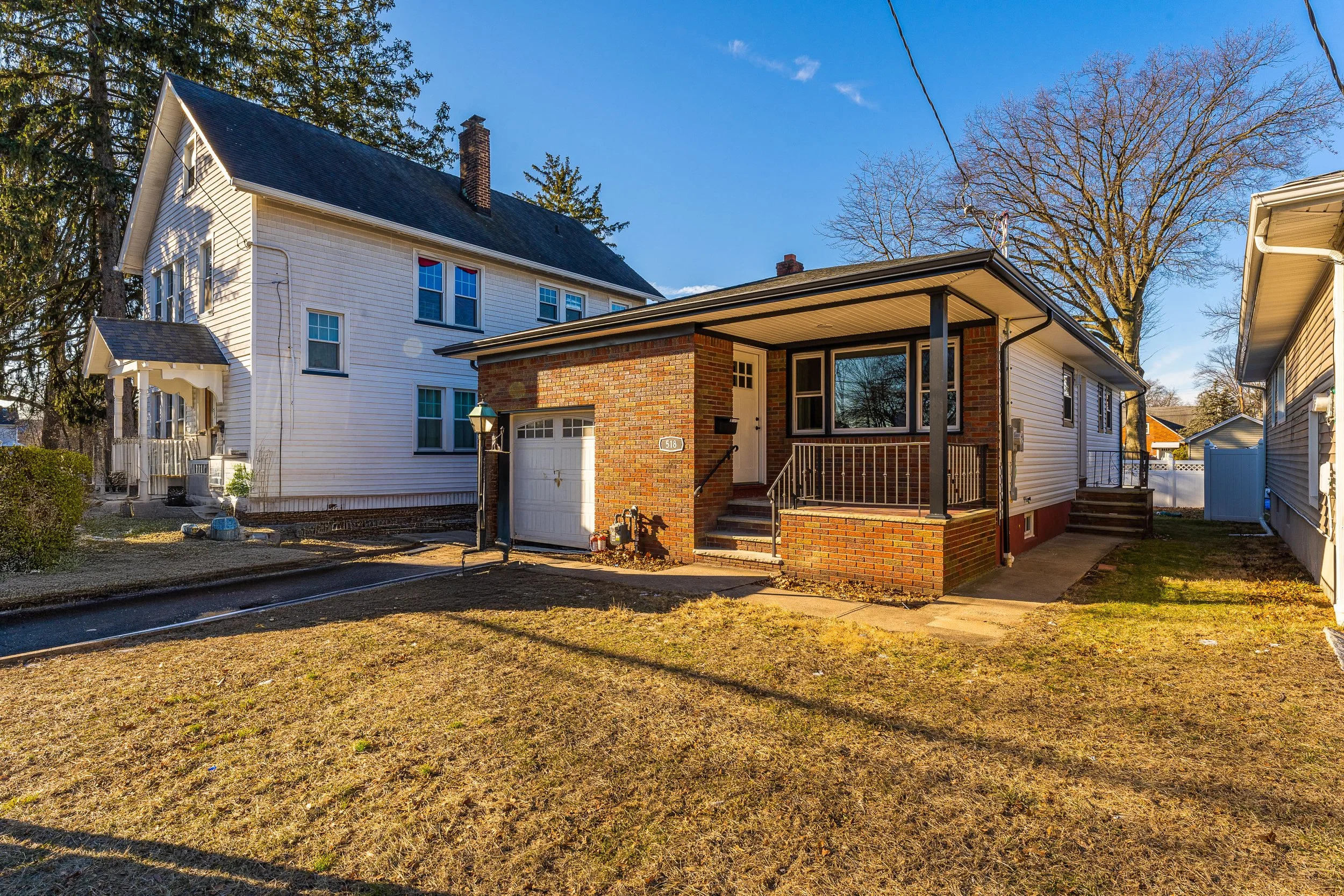 A small house with a brick porch, white siding, and an attached garage, situated in a neighborhood with other houses and large trees, during daytime with clear skies.