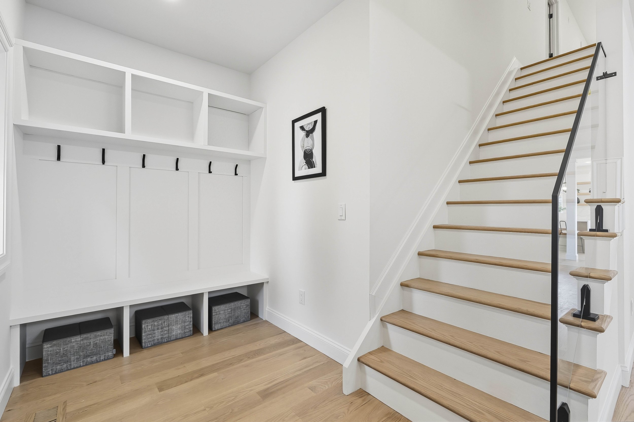 White staircase with wooden steps and black and glass banister, adjacent to white wall with a black and white abstract art print, and white storage bench with cubbies and black and gray storage boxes underneath.