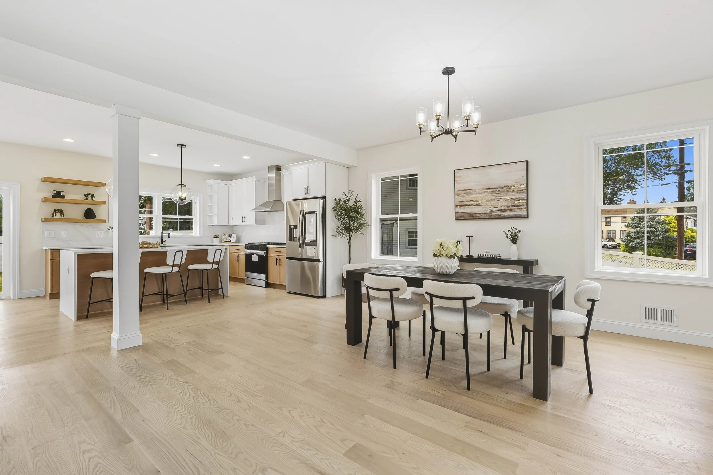 Open-concept kitchen and dining area with light wood flooring, white walls, and modern furniture. The kitchen has white cabinets, a stainless steel refrigerator, and a small bar with two chairs. The dining area features a black table with seven white