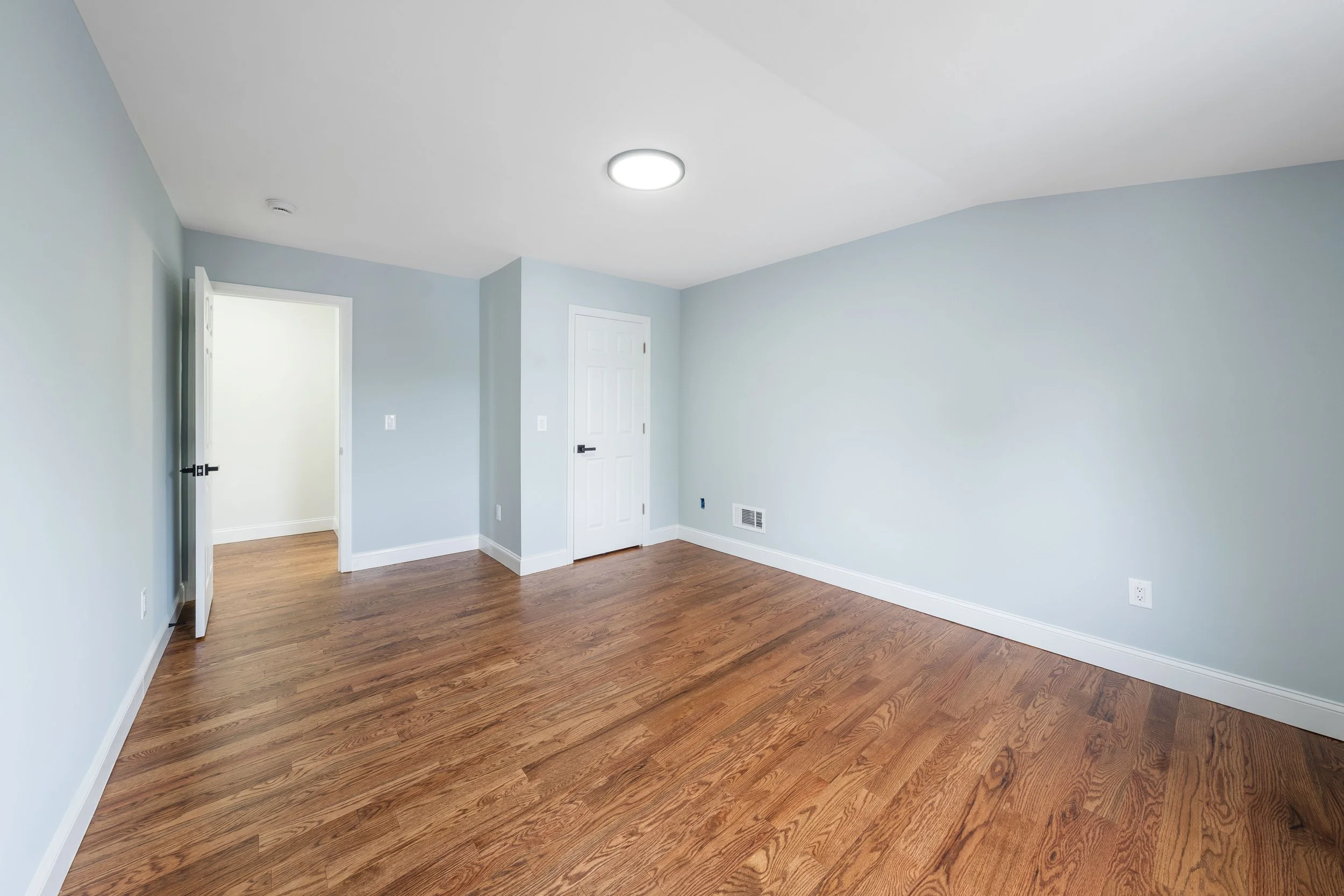 Empty room with light blue walls, hardwood floors, white baseboards, closed white door, and ceiling light.