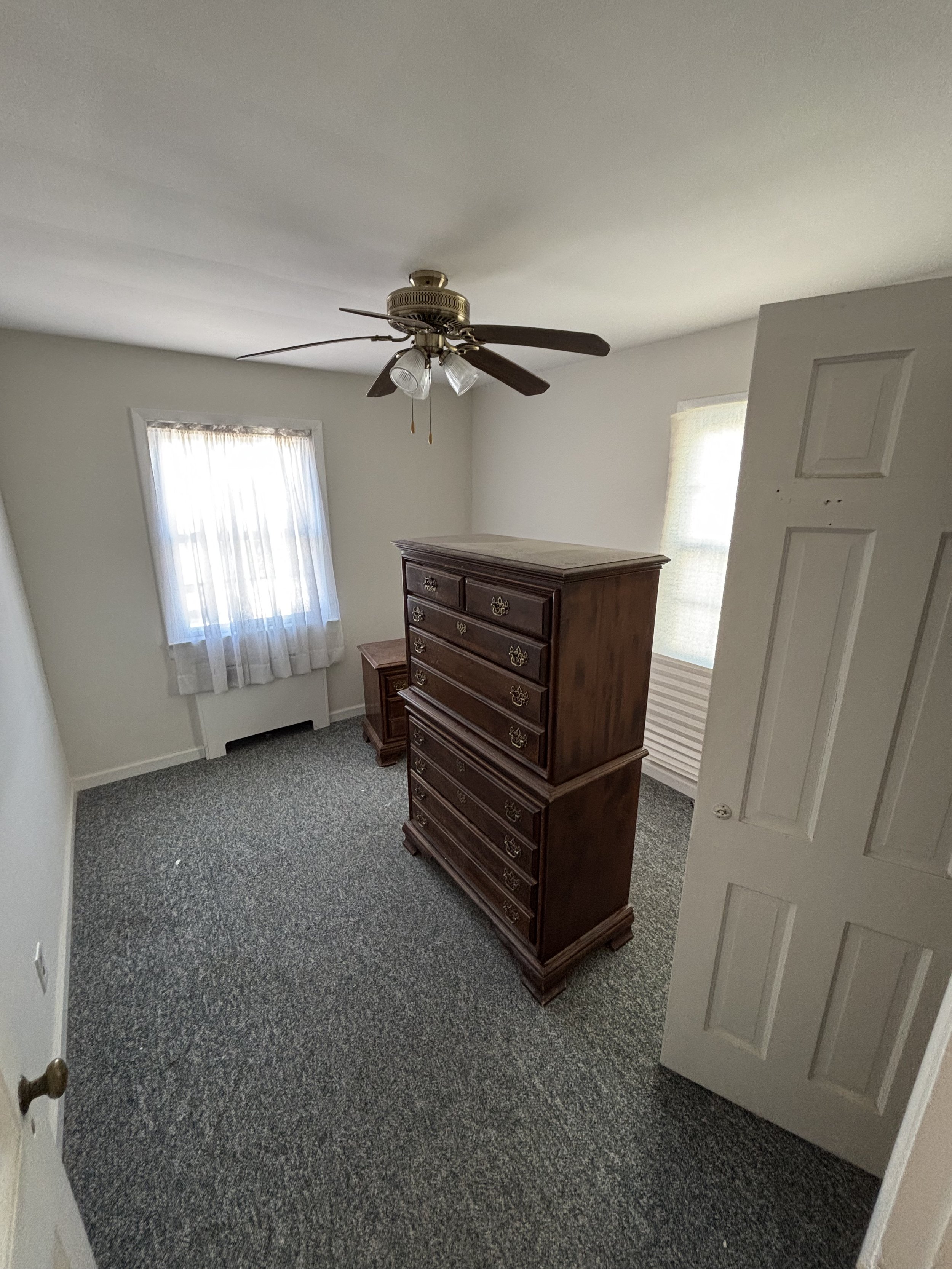 A small bedroom with two windows, one with sheer curtains, a ceiling fan with light fixtures, a wooden dresser, and a door open to the room.