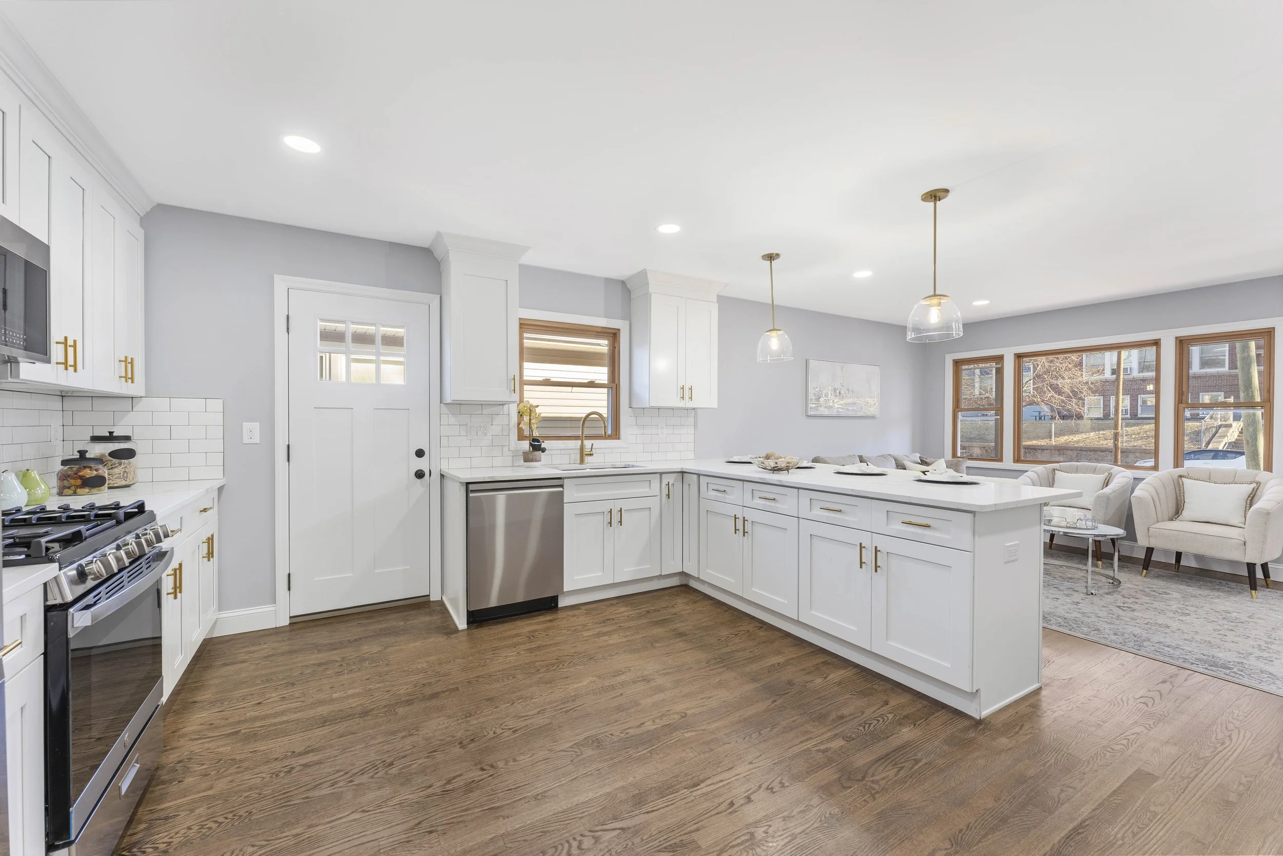 A modern kitchen with white cabinets, a white island, wooden floors, and a living area with large windows and cozy seating.