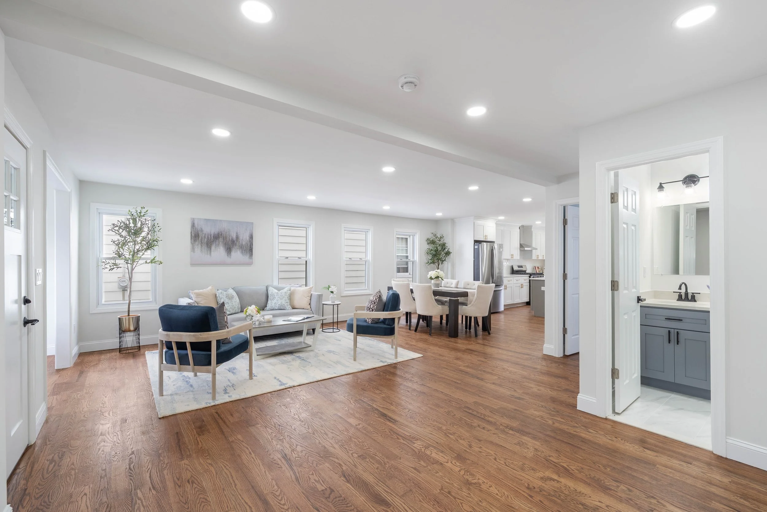 Bright living room with white walls, hardwood floors, white couch, blue armchairs, dining table with chairs, kitchen with white cabinets and stainless steel appliances, small bathroom with gray vanity.