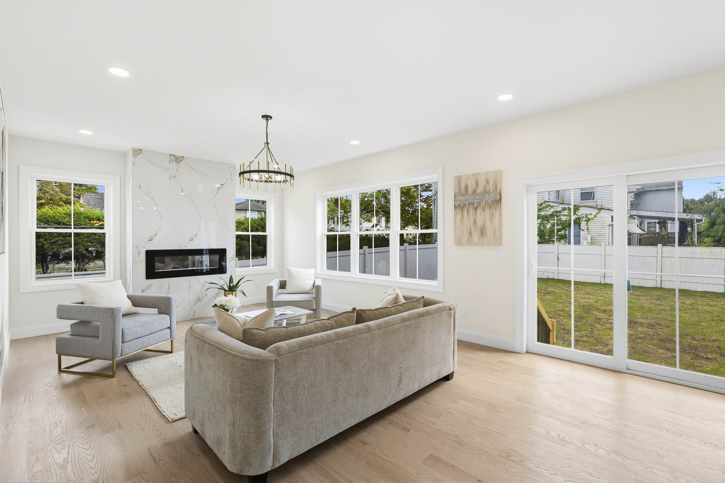 Bright living room with beige sofa, two gray armchairs, a white area rug, large windows, a sliding glass door, a modern fireplace, and a chandelier.