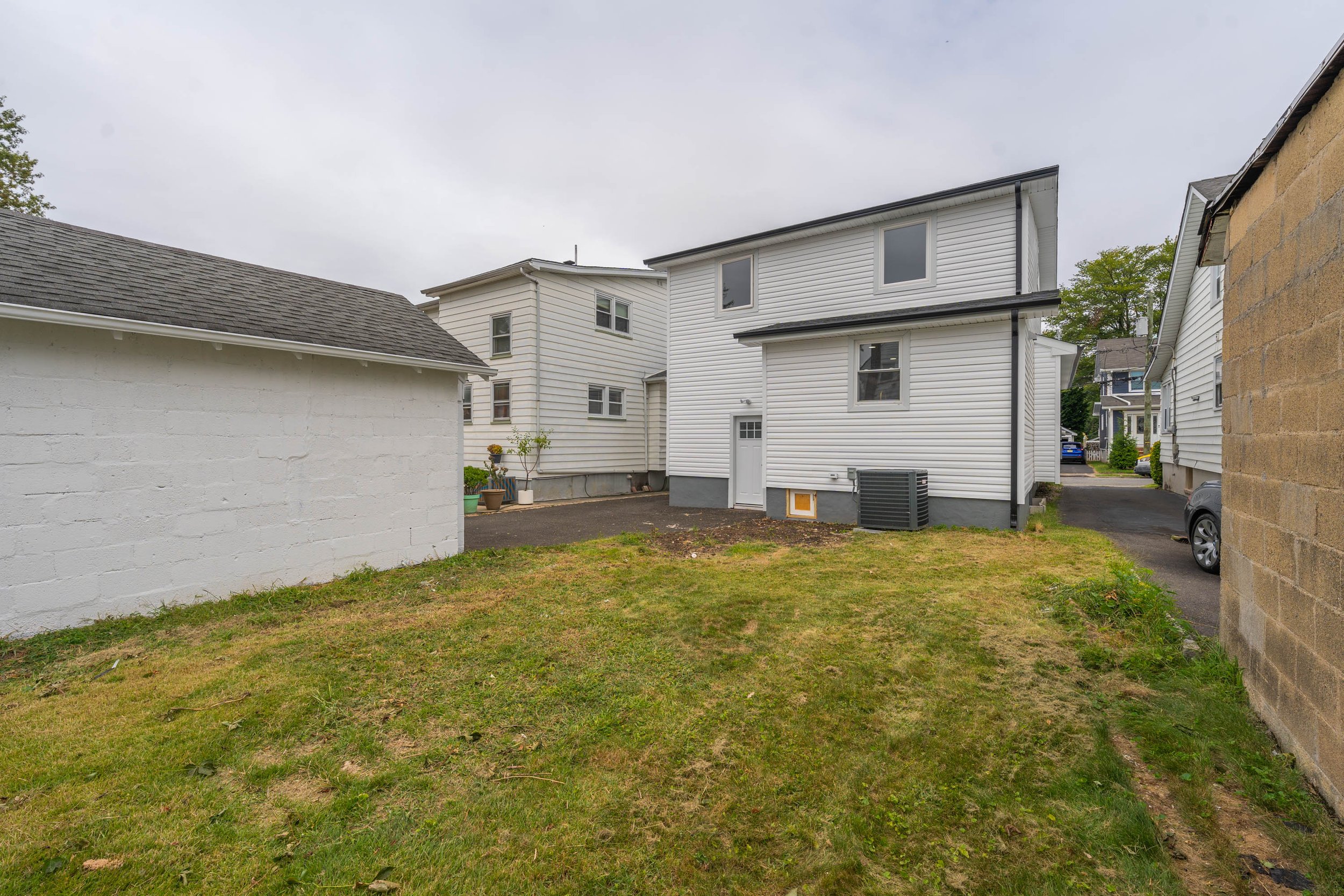 Backyard with a grassy area and a white house with vinyl siding, windows, and a door, surrounded by neighboring houses and a driveway.