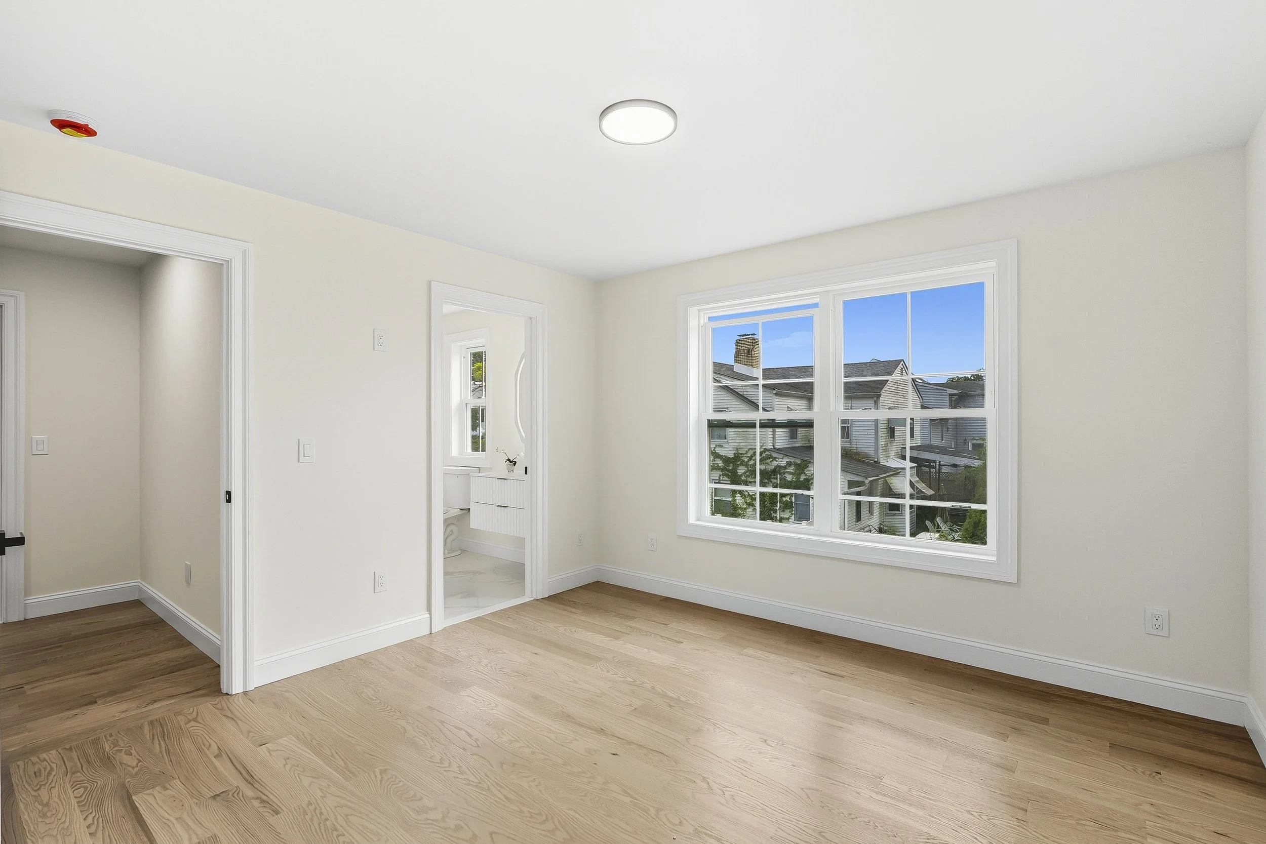Empty room with white walls, large window, light wood flooring, and an en-suite bathroom visible through a door.