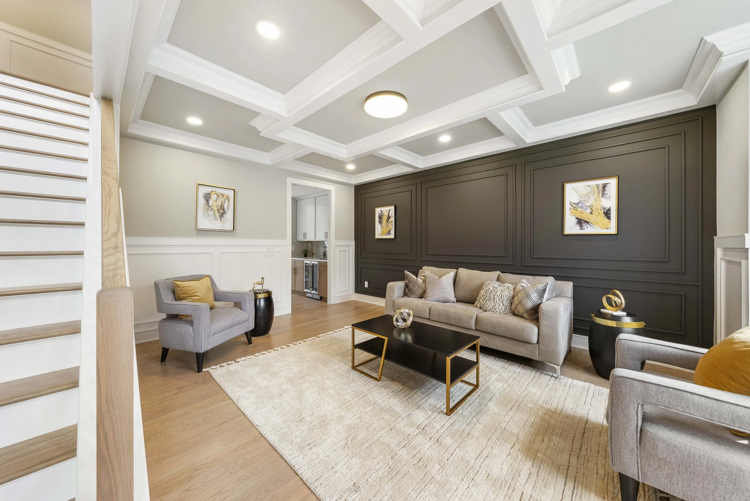 Living room with beige sofas and armchairs, dark accent wall with framed artwork, white coffered ceiling with recessed lighting, light wood flooring, black and gold coffee table, side tables, and decorative sculptures
