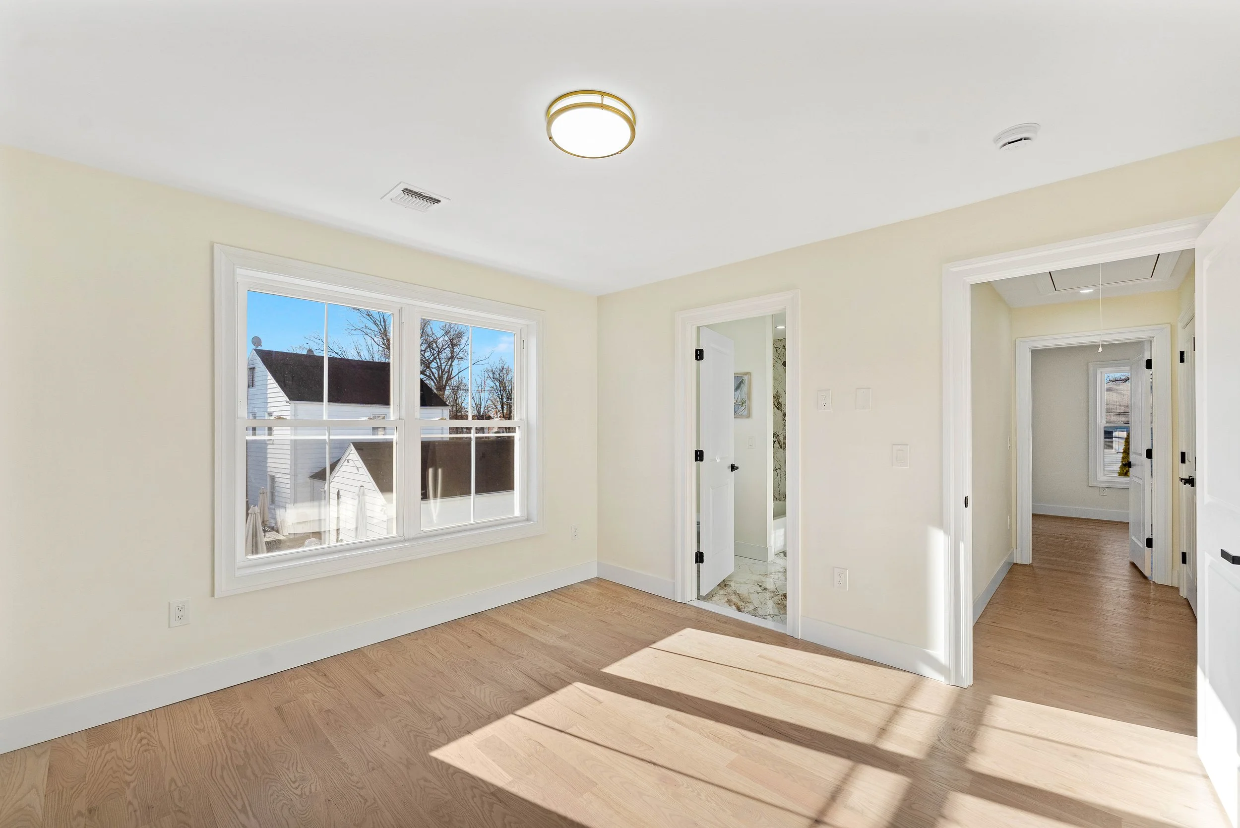 Empty room with large window, light wood floors, white walls, and doors leading to other rooms. Sunlight casts shadows on the floor.