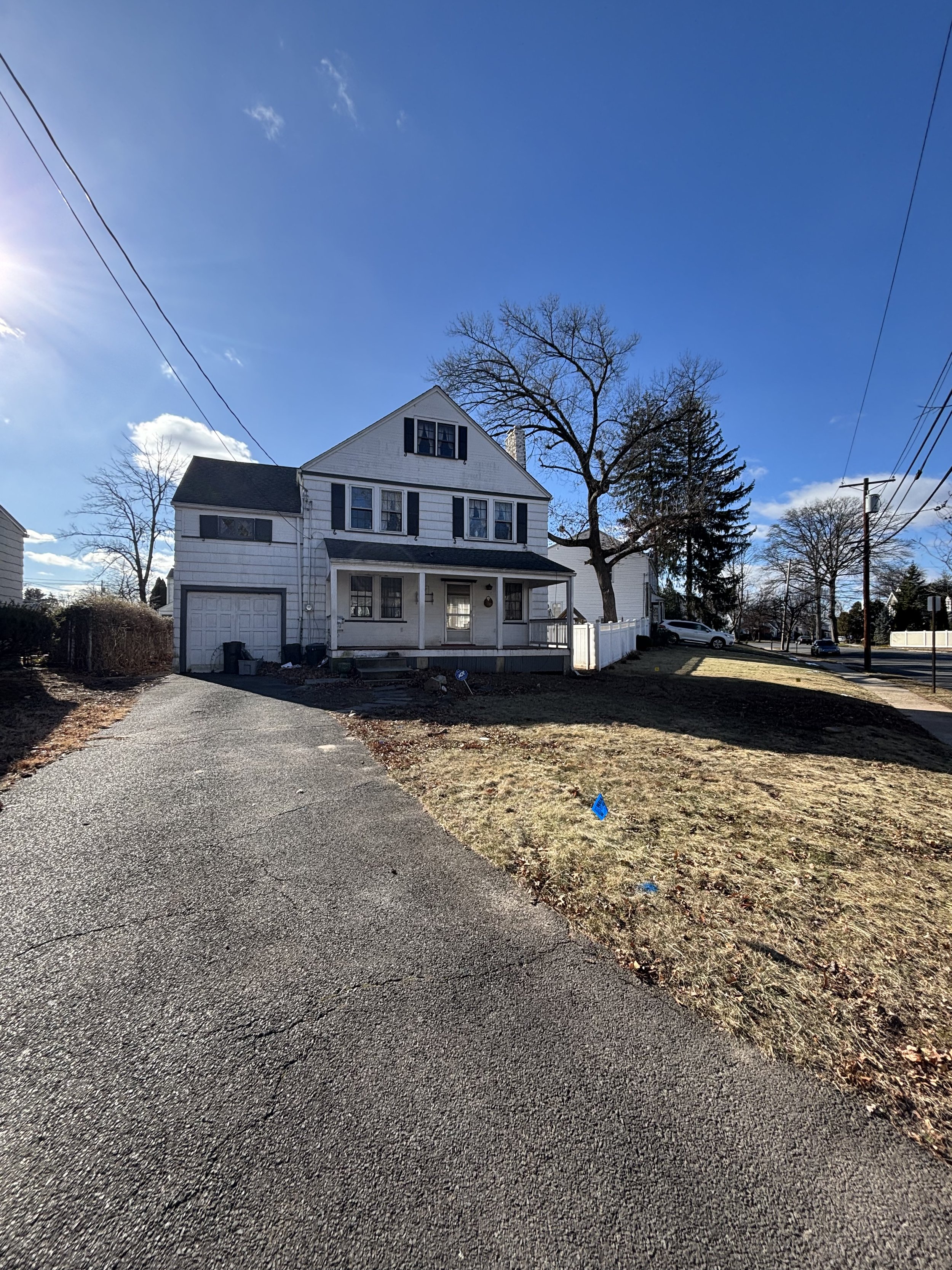 A two-story white house with black shutters and a front porch, situated on a lawn with a driveway leading up to an attached garage, under a clear blue sky with some trees and power lines.