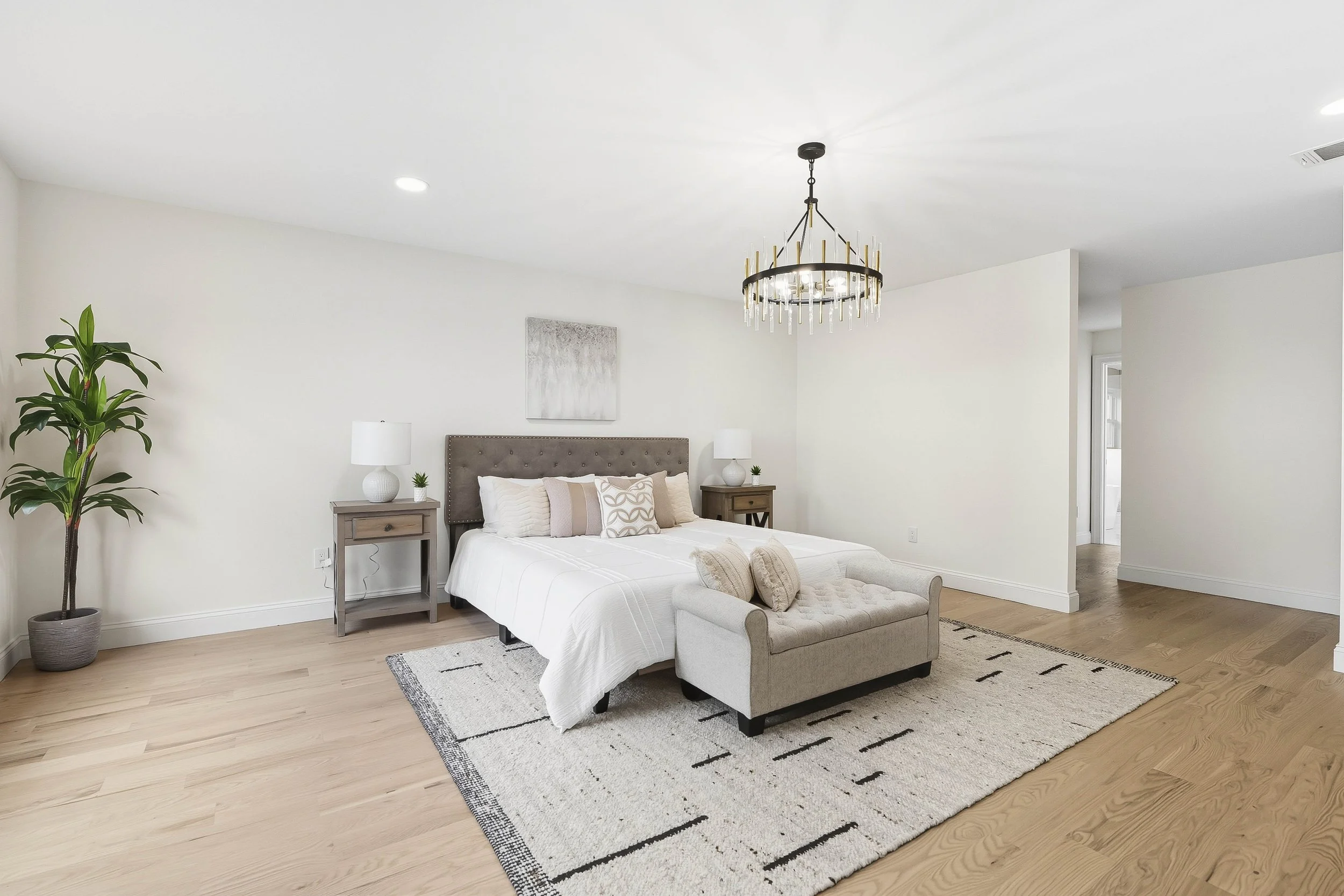 A modern bedroom with a large bed, gray upholstered headboard, white bedding, and decorative pillows. Flanking the bed are matching wooden nightstands with white lamps and small plants. A tufted bench sits at the foot of the bed. The room features wo