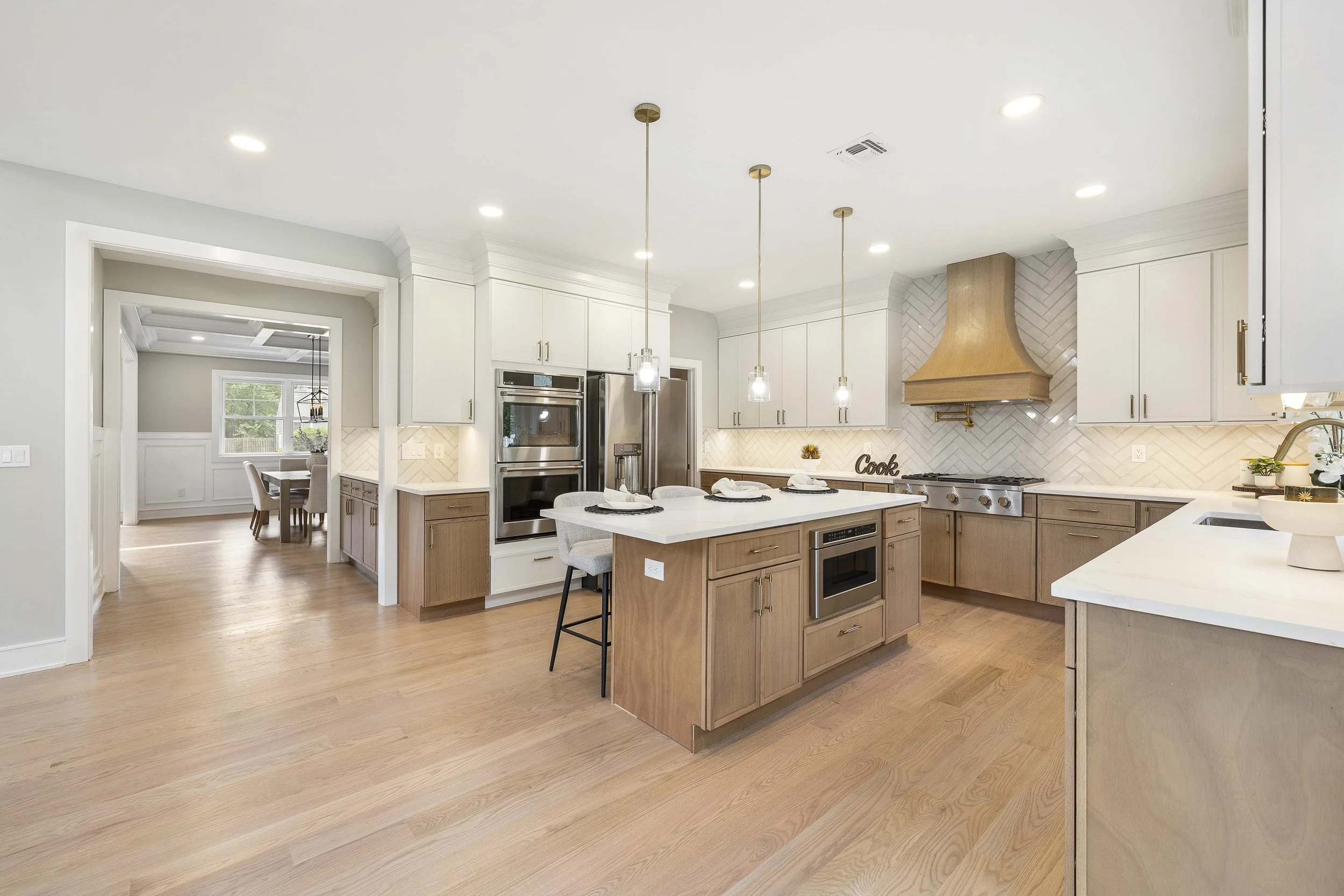 Modern kitchen with white cabinets, wooden accents, and a central island with bar stools. Stainless steel appliances, pendant lighting, and a herringbone backsplash behind the stove. Adjacent dining area visible through an open doorway.